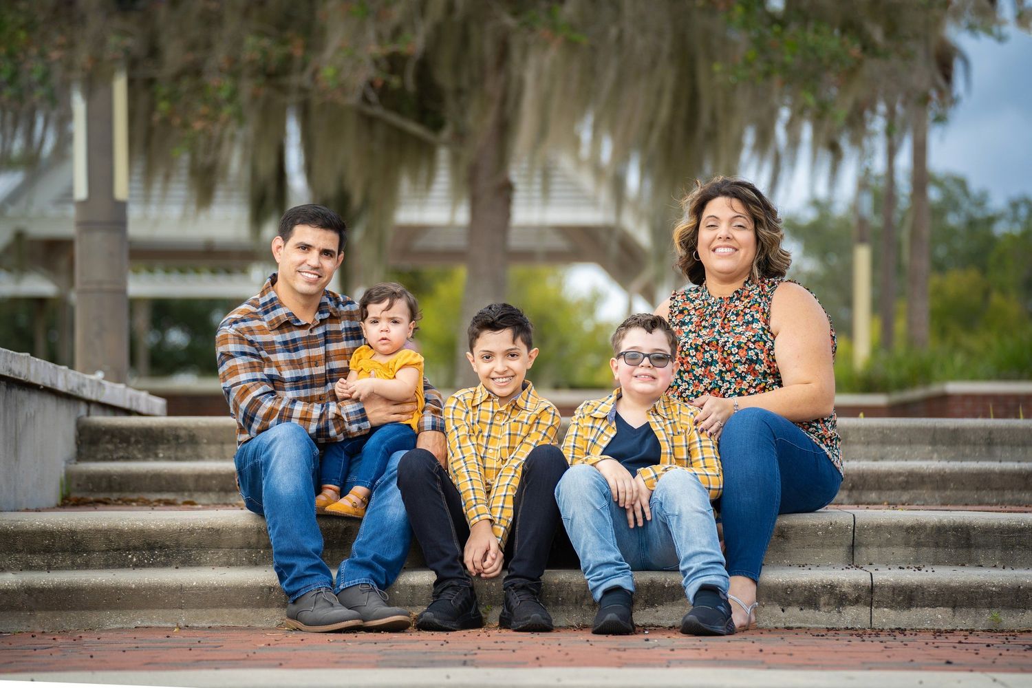 A family of four sits casually on outdoor stone steps with Spanish moss draped trees in the background.