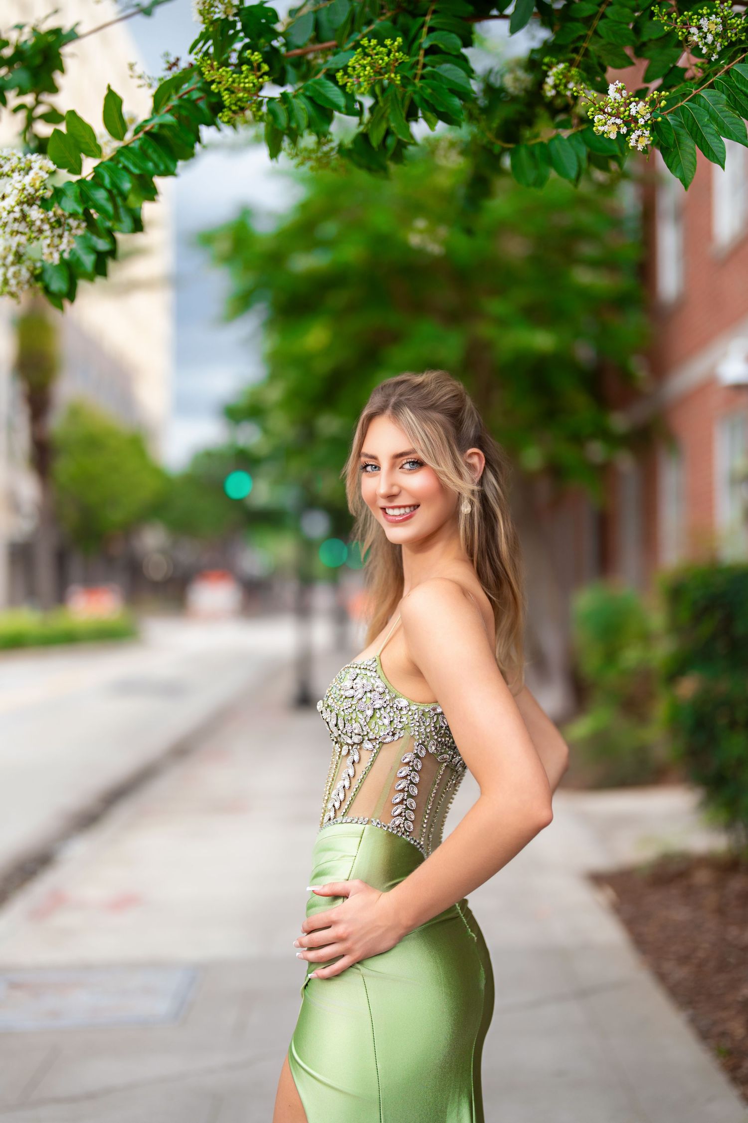 Young woman in elegant green embellished strapless gown posing on urban sidewalk beneath flowering trees.