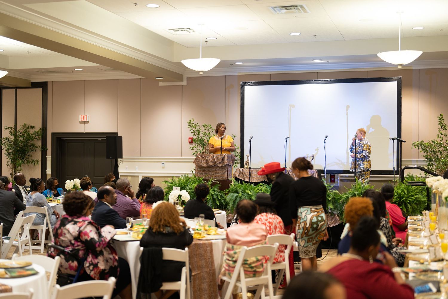 Guests seated at banquet tables listen to speakers at a formal indoor event in a decorated ballroom.