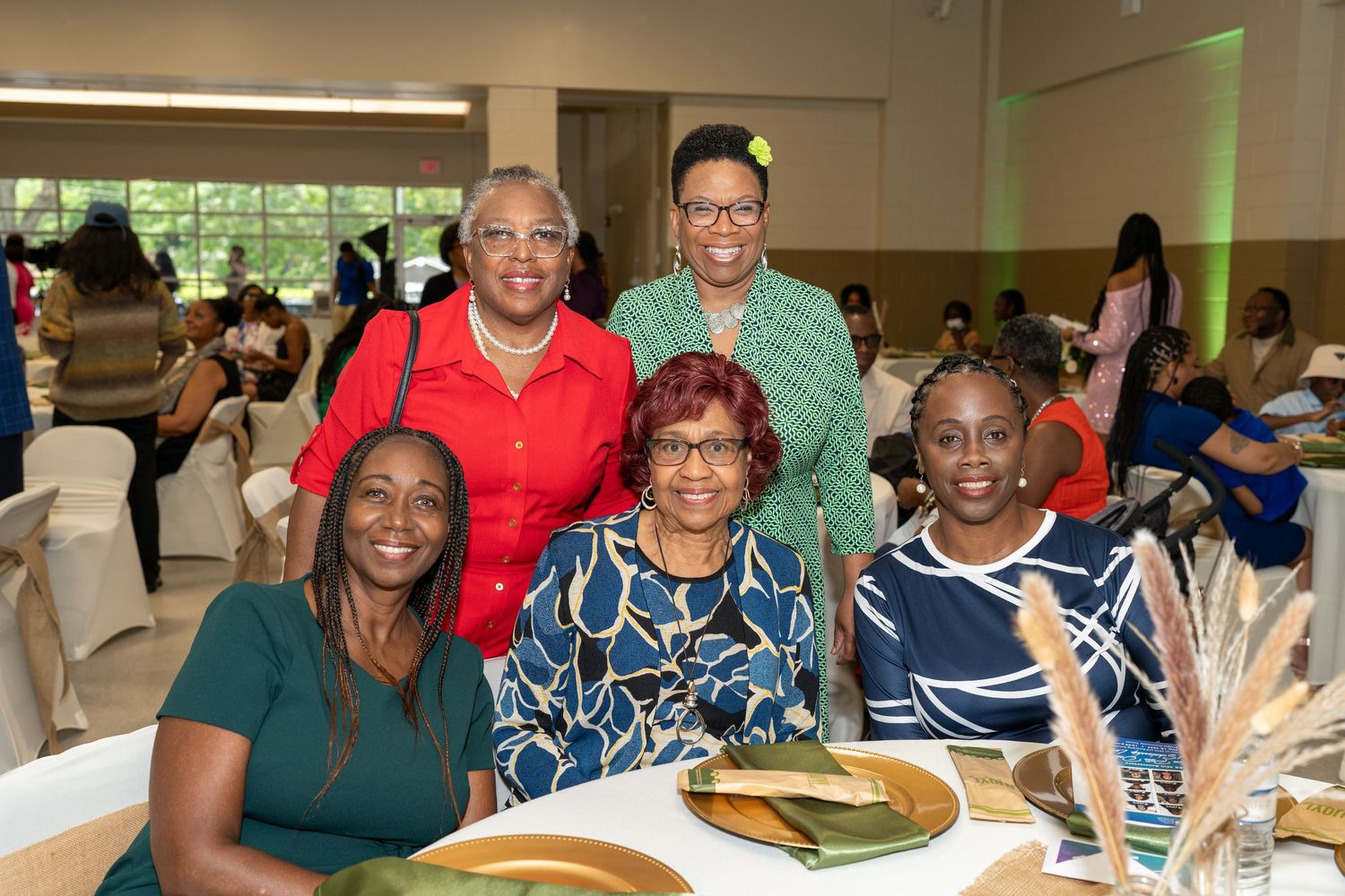 Five women smiling together at a banquet event, seated and standing around a decorated table.