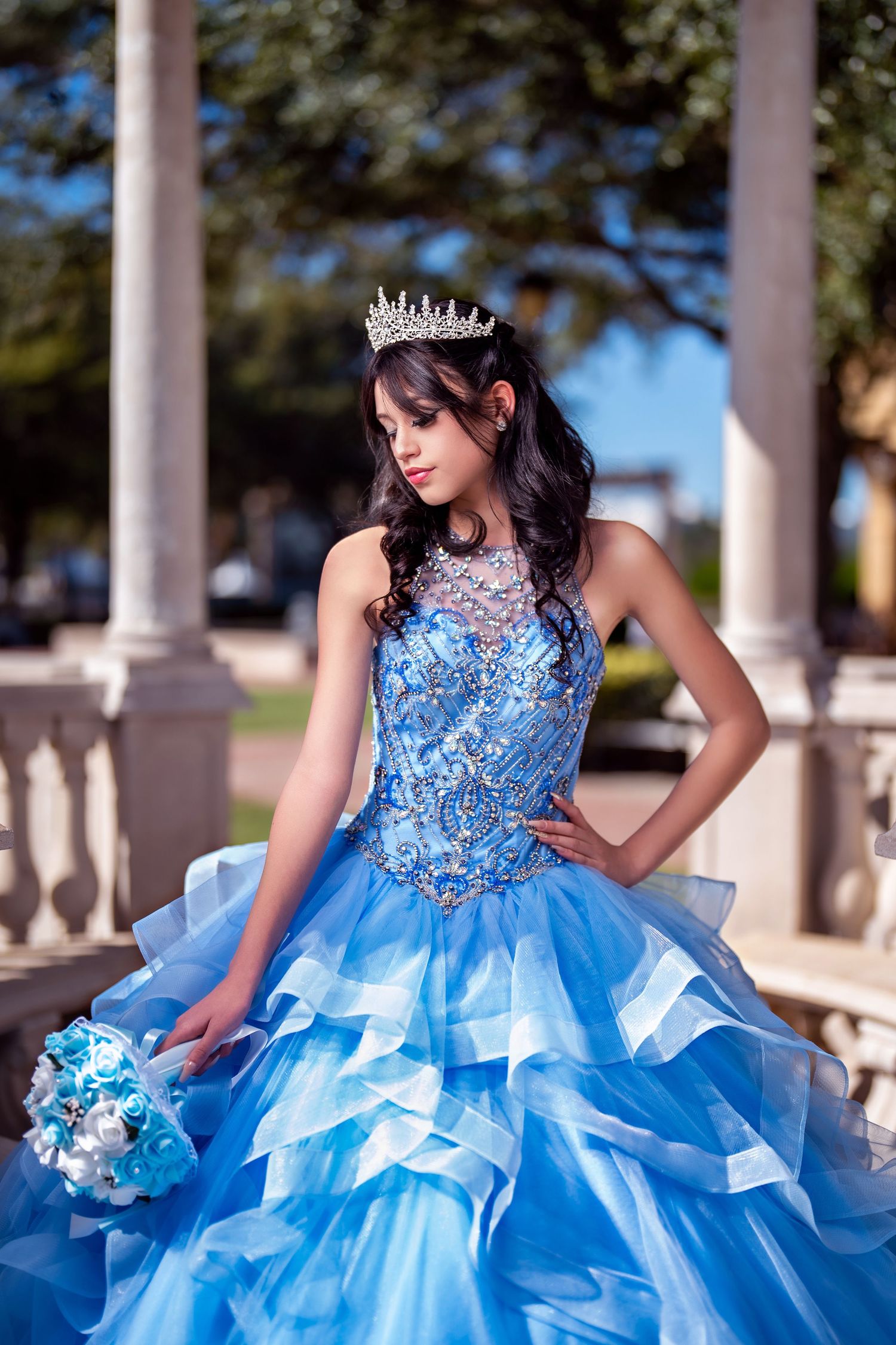 Young Latina girl in blue ball gown with silver tiara holds bouquet at quinceañera celebration outdoors.