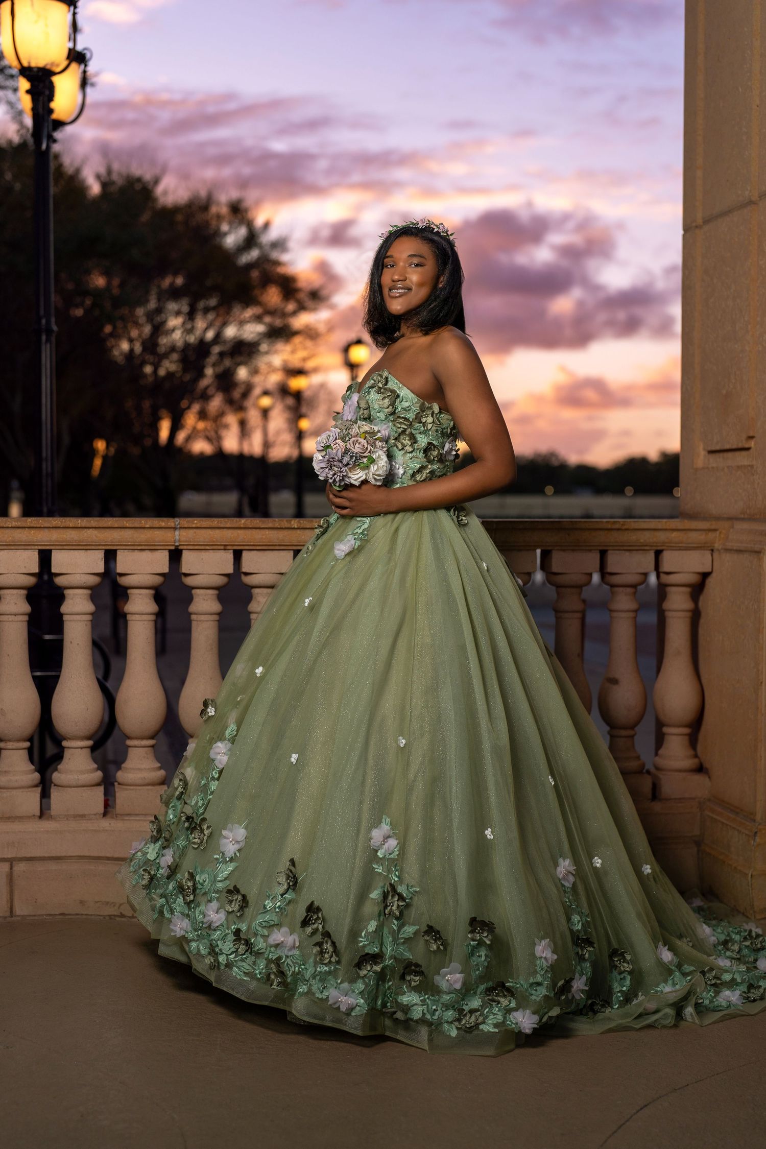 Young woman in sage green floral ball gown with tiara poses on balcony at sunset during quinceañera celebration.