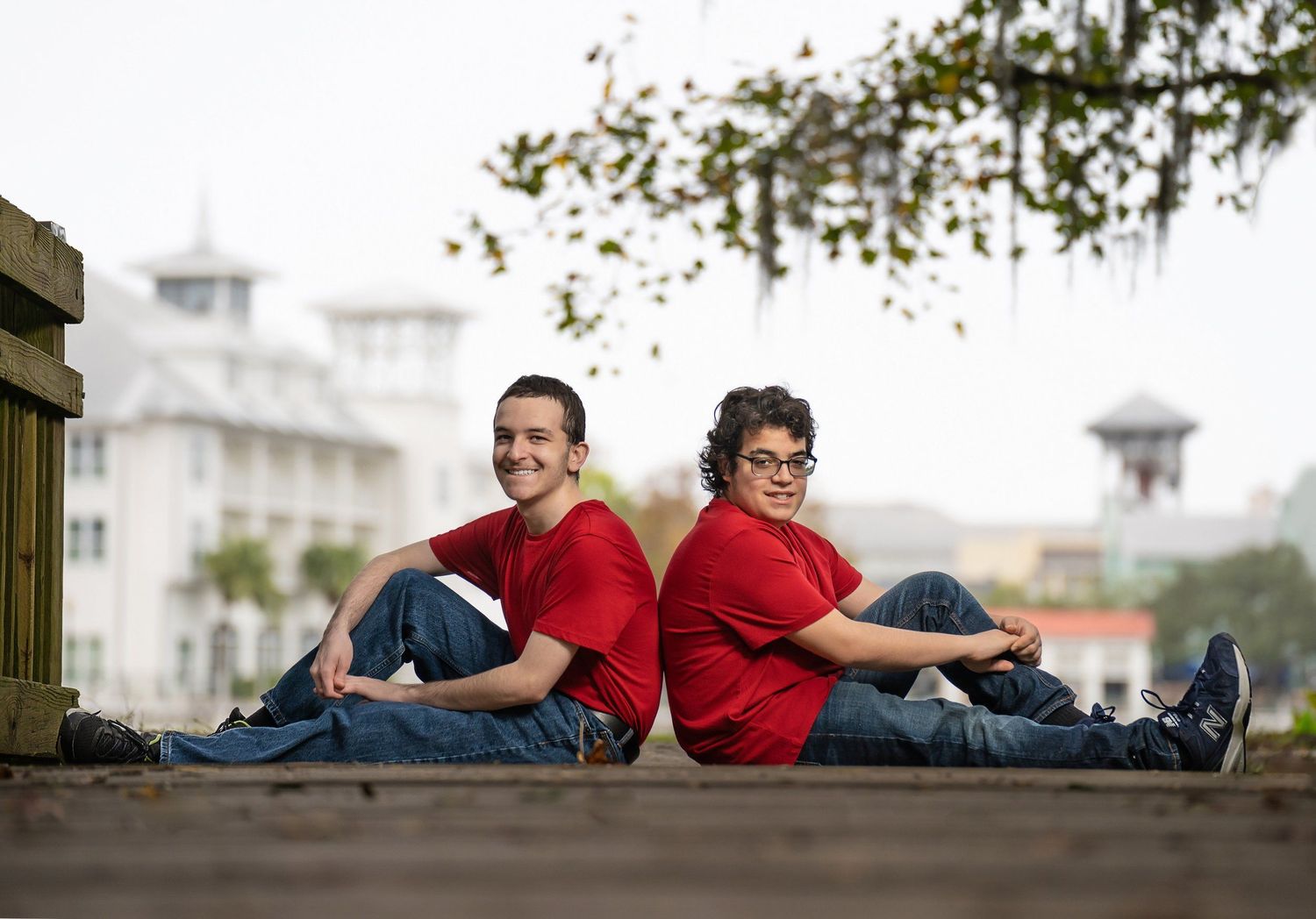 Two people in matching red shirts sit back-to-back on wooden steps outdoors, smiling, with a white building in the background.