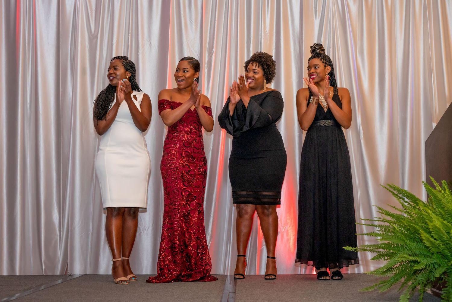 Four women in elegant formal gowns applauding on stage at a glamorous event against a white draped backdrop.