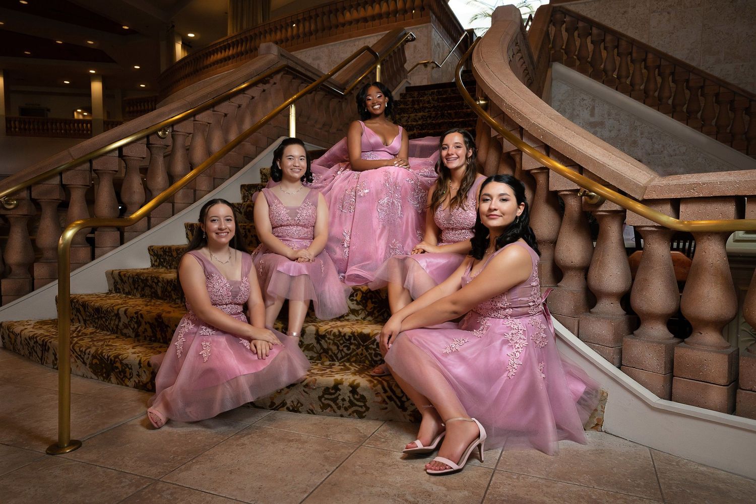 Five women in matching pink tulle dresses pose elegantly on a grand staircase for a formal event.