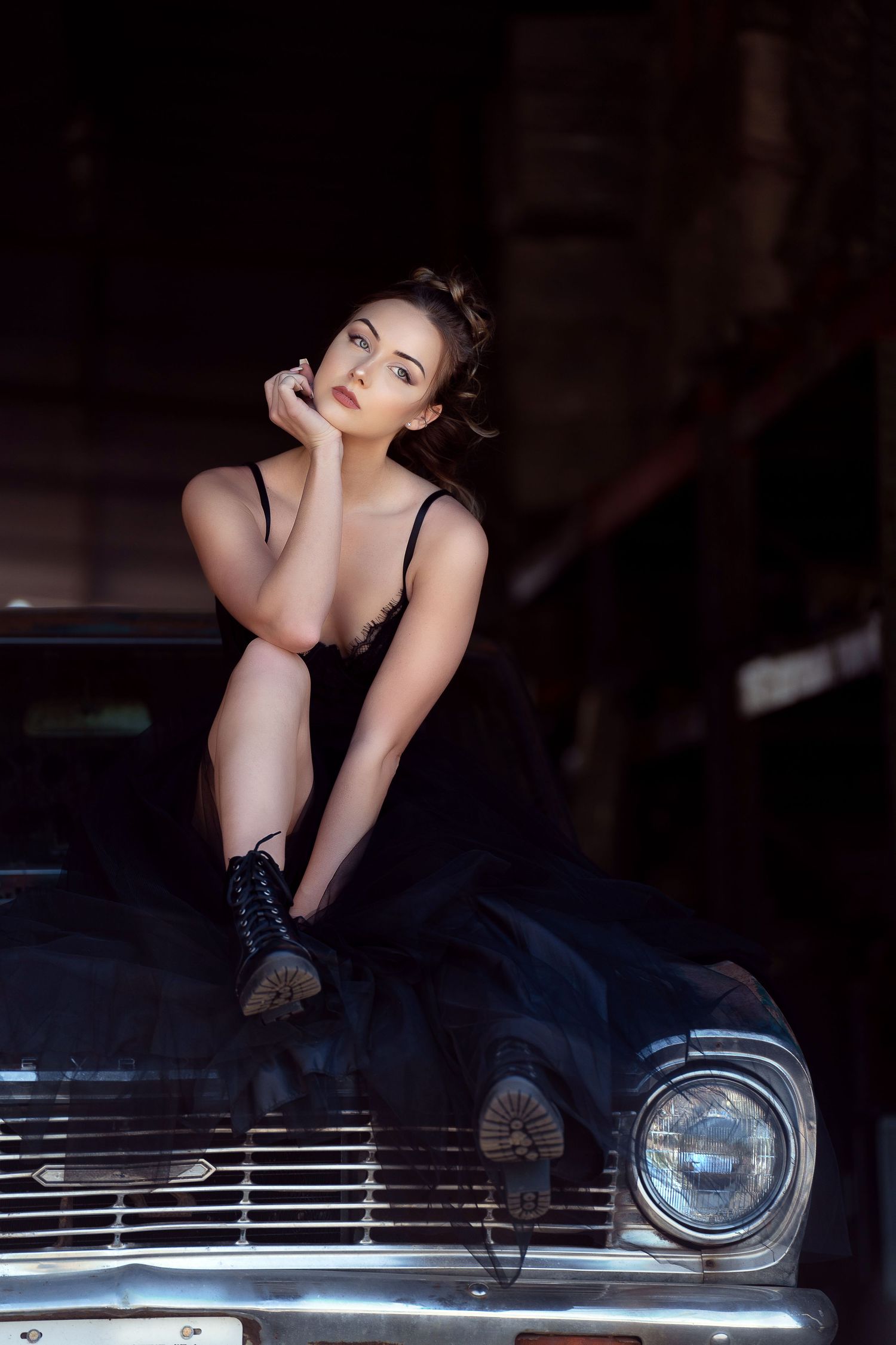 Young woman in black dress poses on vintage car hood in dark garage, looking up thoughtfully.