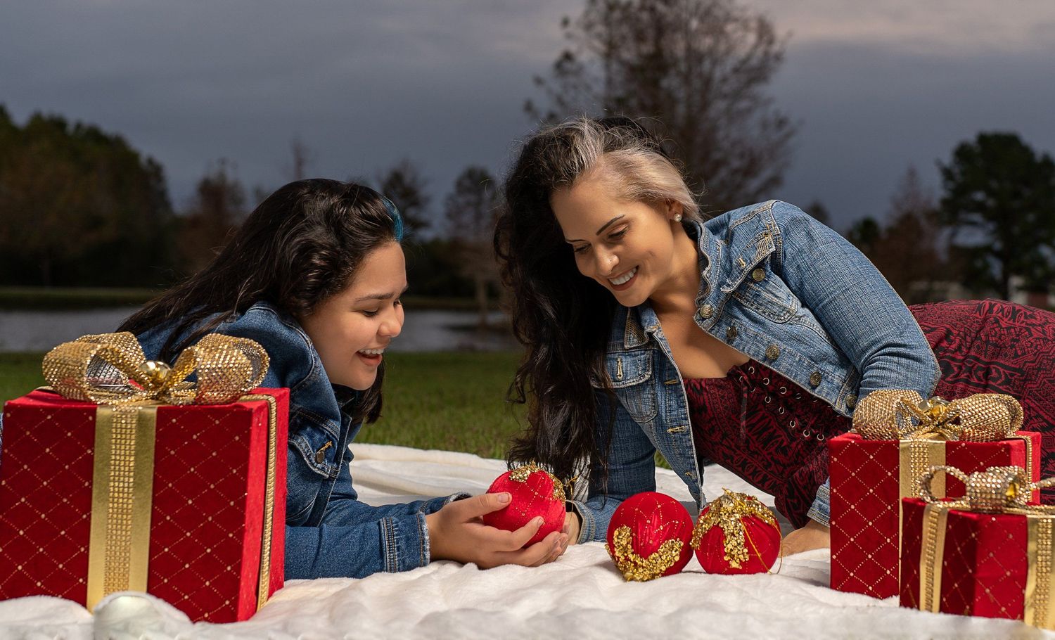 Two people in denim jackets sit on a white blanket surrounded by wrapped red and gold Christmas presents outdoors.
