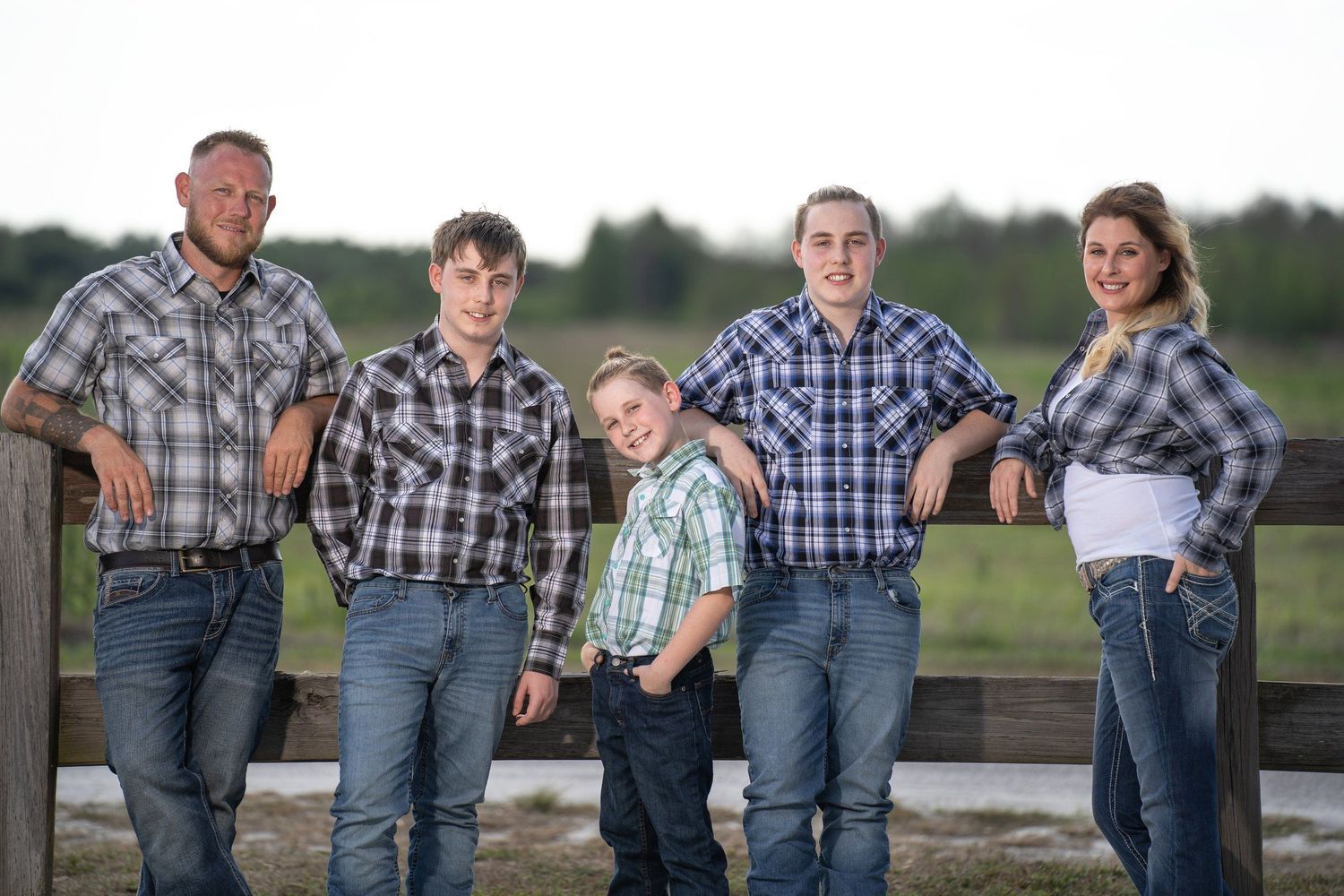 A family in coordinating plaid shirts poses together along a wooden fence at dusk.