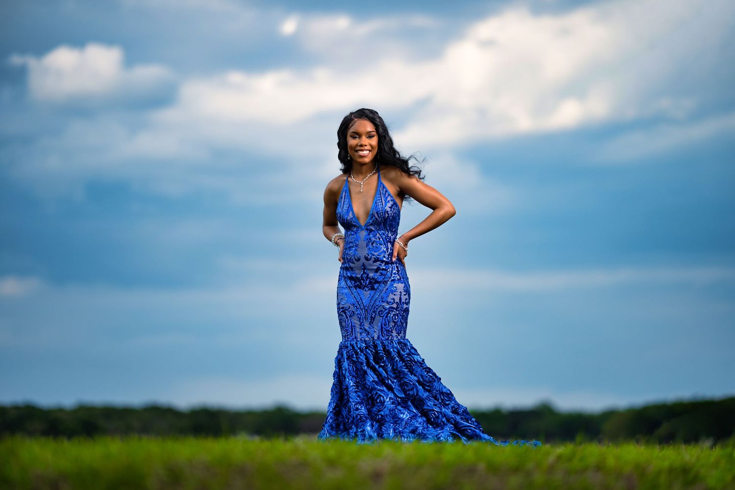 A young Black woman in a royal blue sequin mermaid gown poses gracefully in a green field under a dramatic cloudy sky.