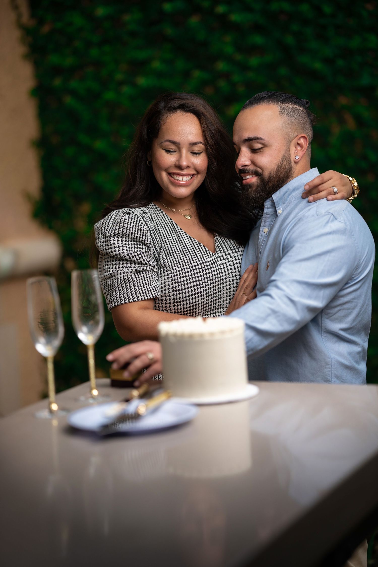 Couple embracing and smiling beside a white cake and champagne glasses against a green ivy wall.