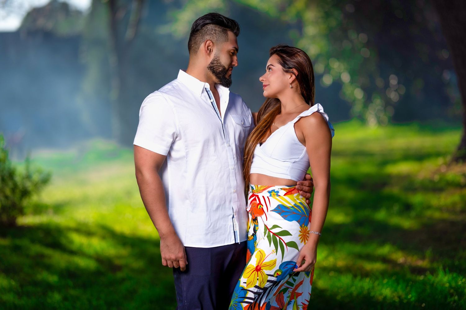 Couple gazing at each other in a lush green park, man in white shirt, woman in floral skirt.