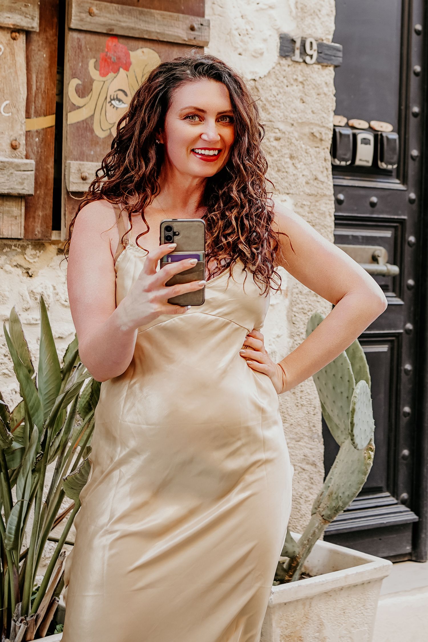 Cristina Nicole Chief Marketing Officer in beige sleeveless dress poses with phone against stone wall with green plants in background.