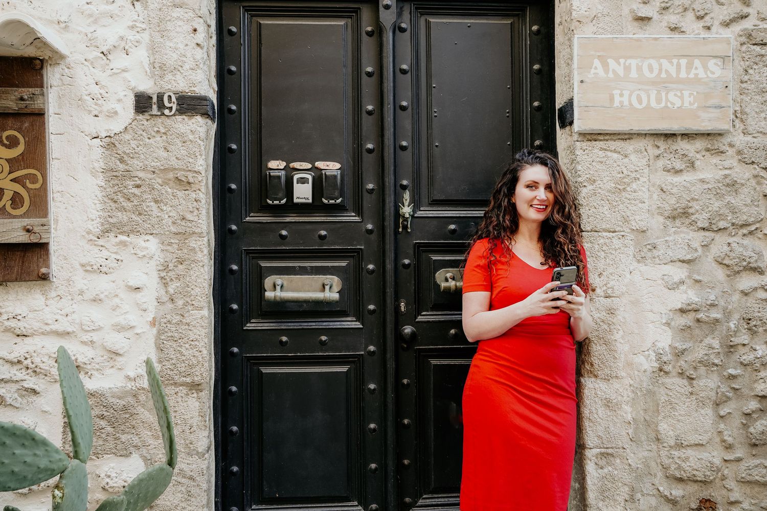 Cristina Nicole in vibrant red dress standing against rustic stone wall with black double doors in Mediterranean setting.