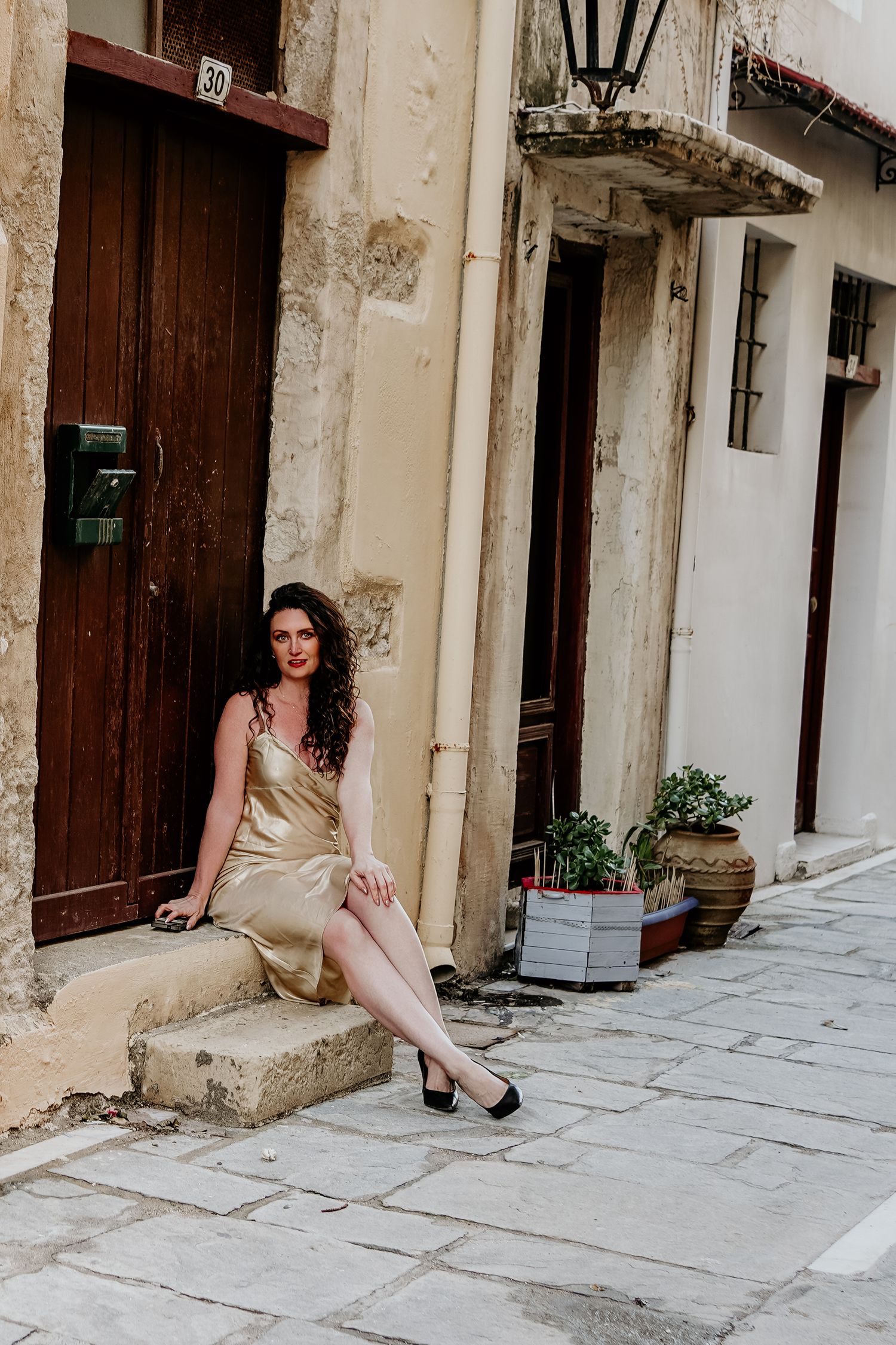 Cristina Nicole Chief Marketing O in a beige dress sits on stone steps in a rustic Mediterranean alleyway with white walls and wooden doors.