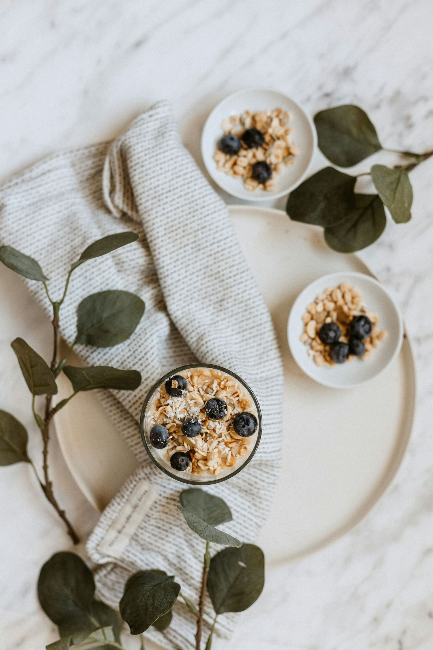Overhead view of yogurt parfaits with granola and blueberries on marble surface with eucalyptus leaves and white linen napkin.