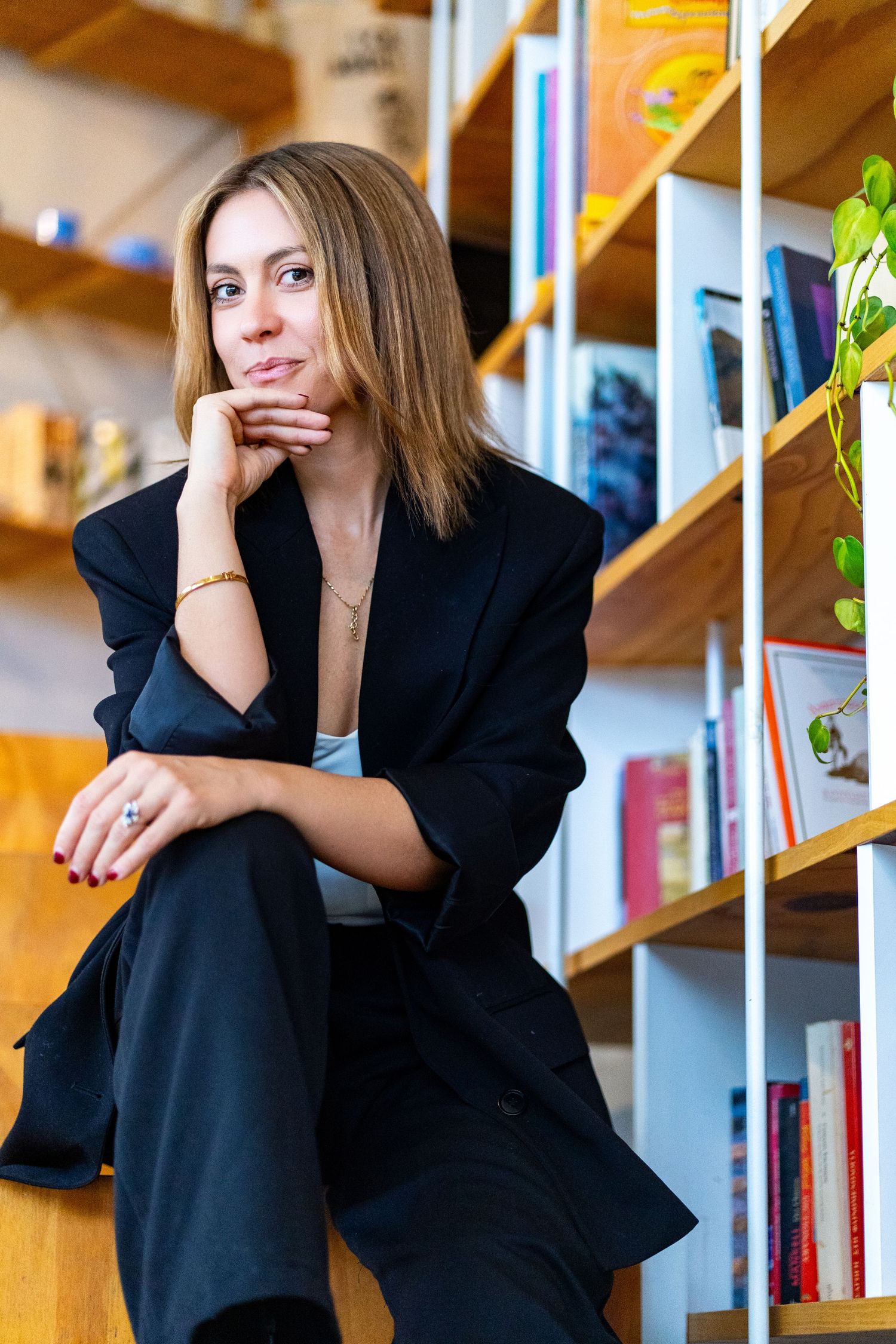 Professional wearing black business suit sits thoughtfully by wooden bookshelf in modern office setting.
