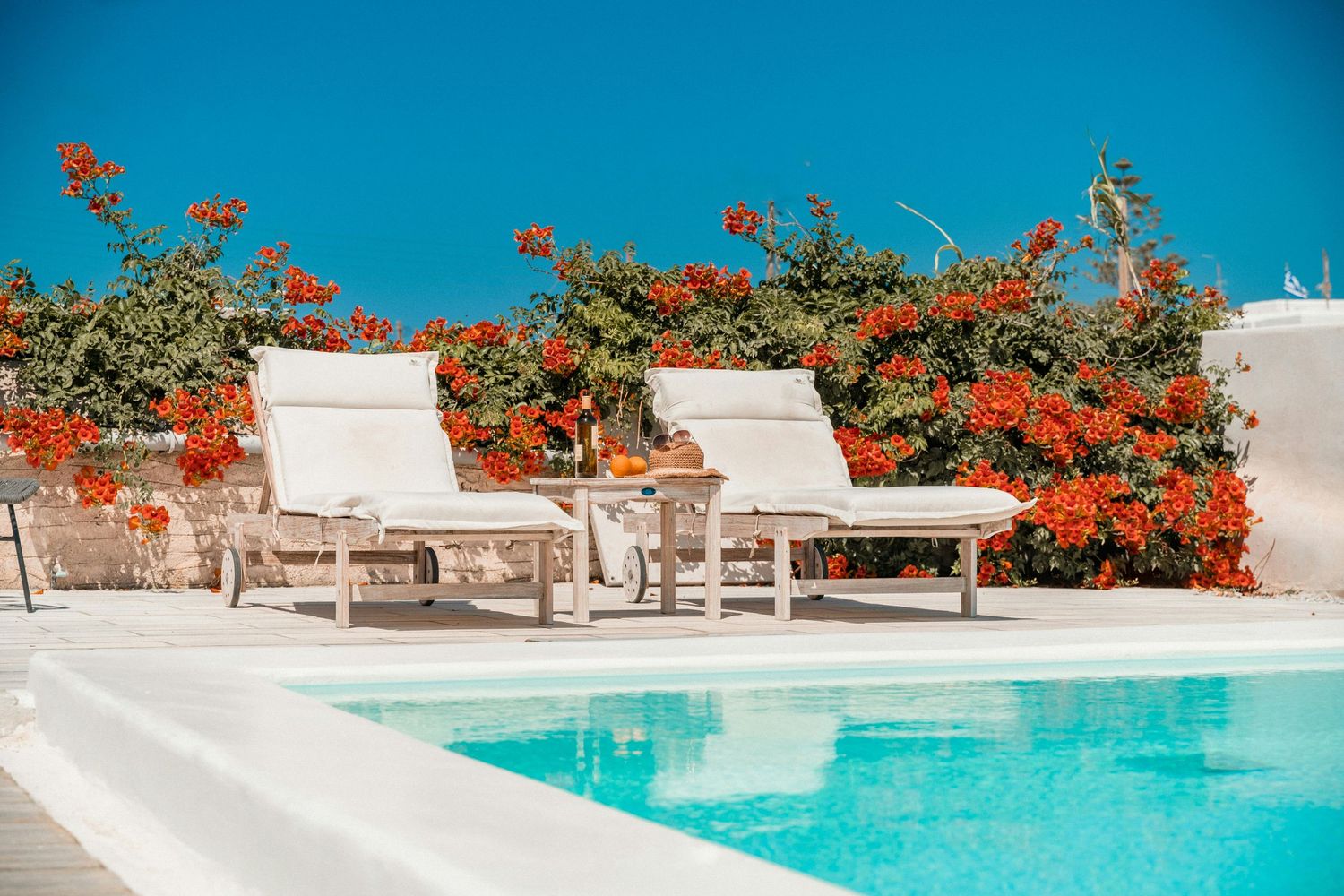 White lounge chairs beside turquoise swimming pool with vibrant orange bougainvillea flowers against blue sky.