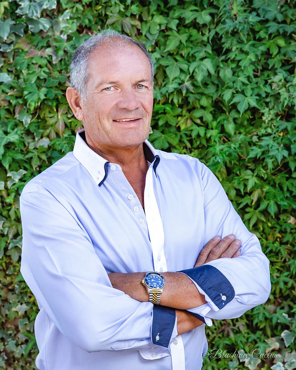 Person in white button-down shirt poses against green foliage background with crossed arms and warm smile.