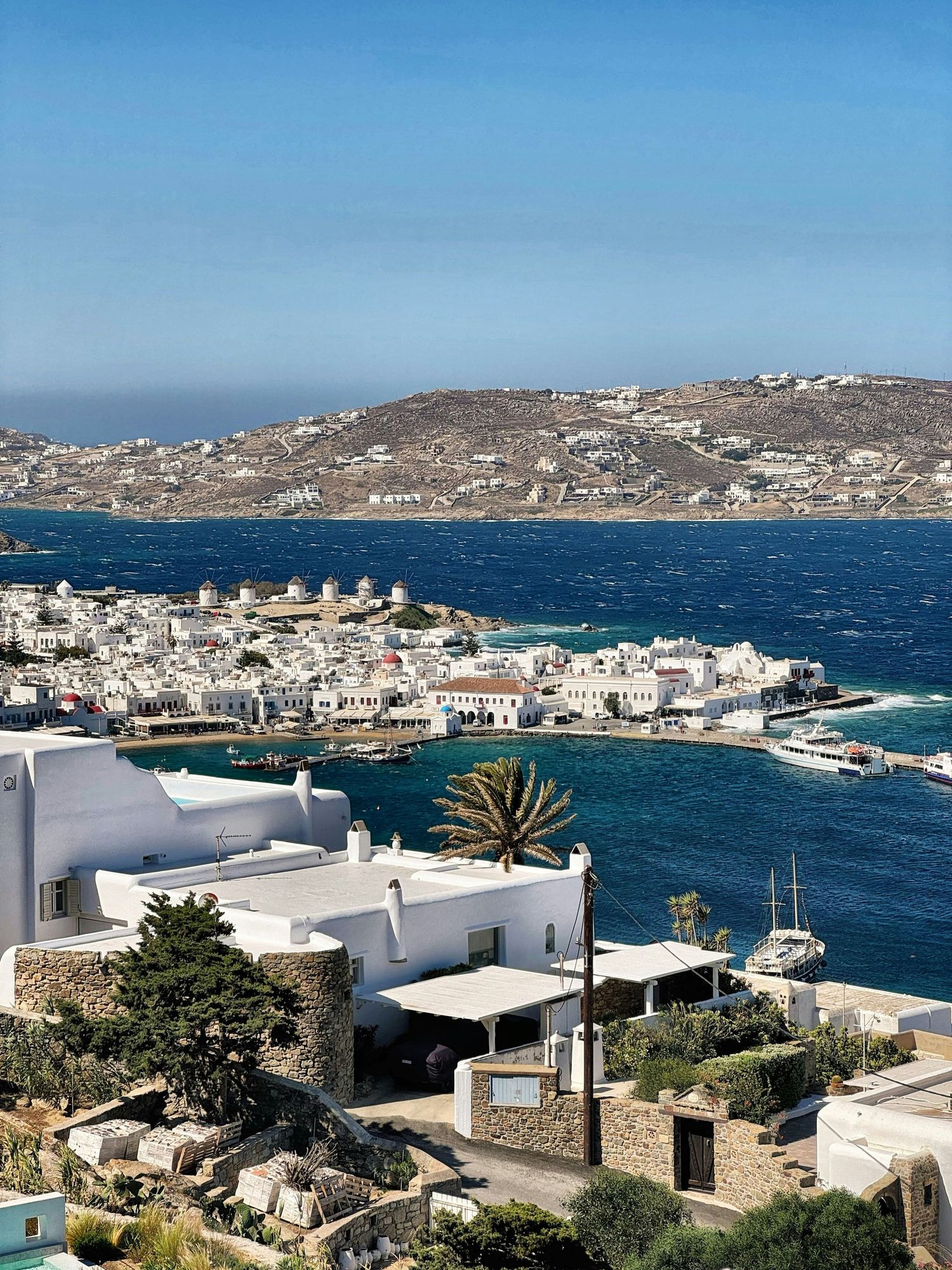 Panoramic views of white buildings and windmills along the coastline of Mykonos, Greece with blue Mediterranean waters.