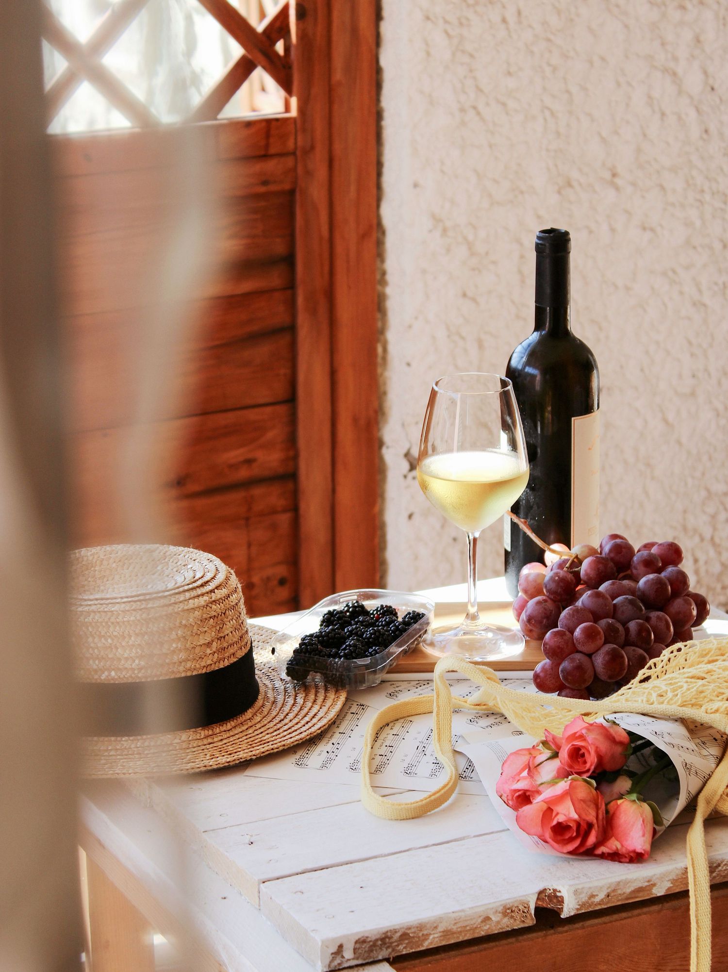 Rustic table setting with wine bottles, grapes, and straw hat in warm sunlight creating an inviting Mediterranean atmosphere.