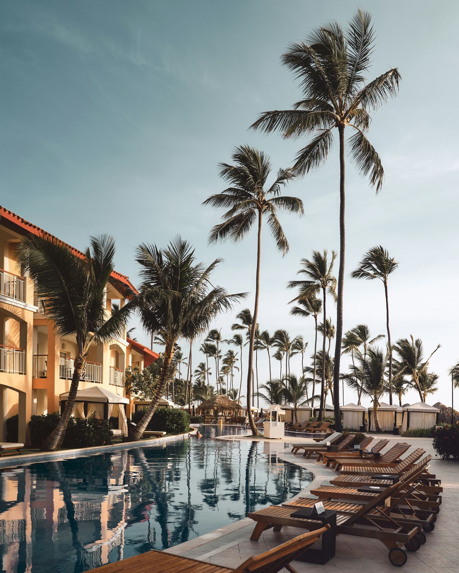 Luxury beachfront resort with infinity pool surrounded by palm trees and lounge chairs at sunset.