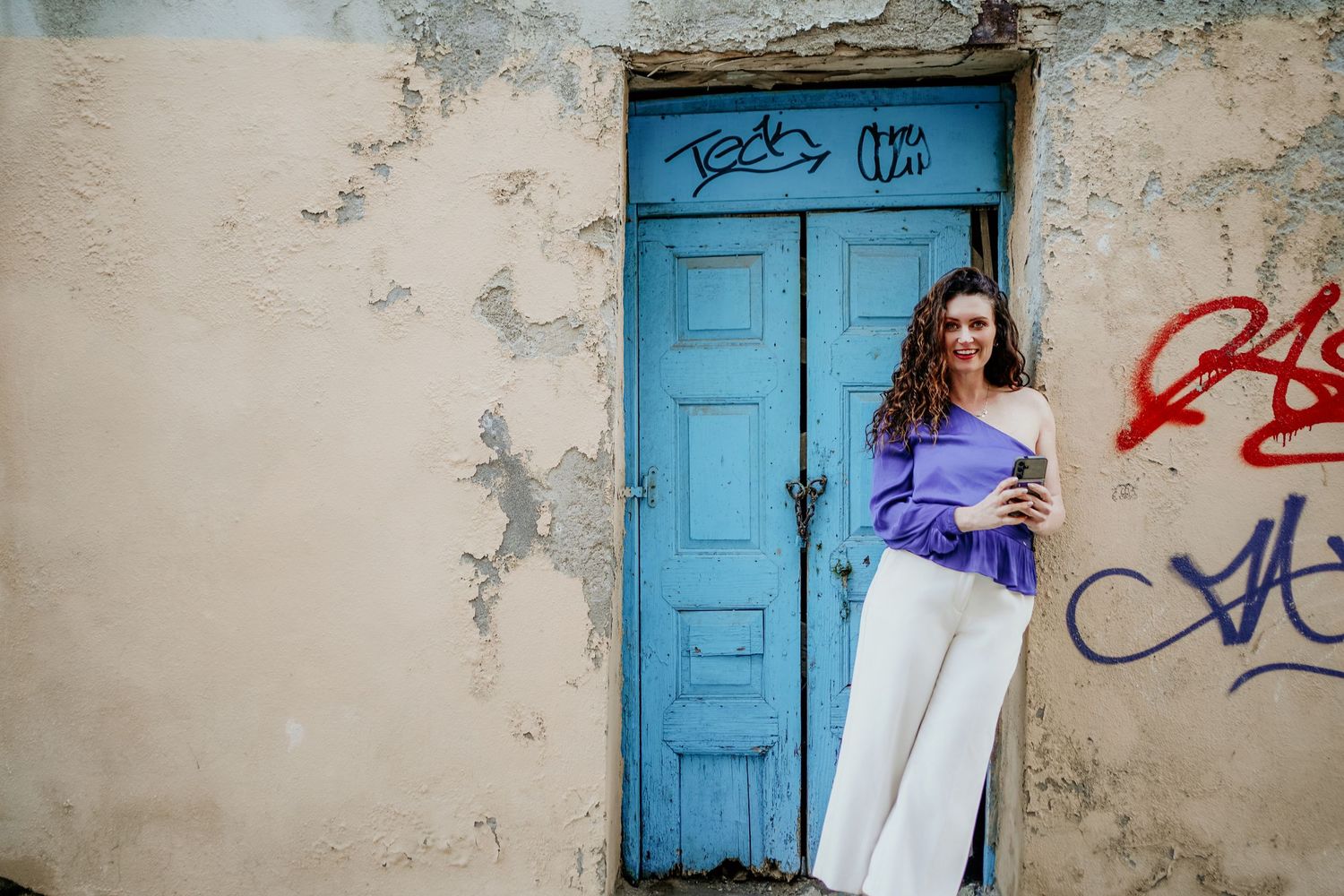 A bright turquoise door frames a casual pose against a weathered wall with graffiti.