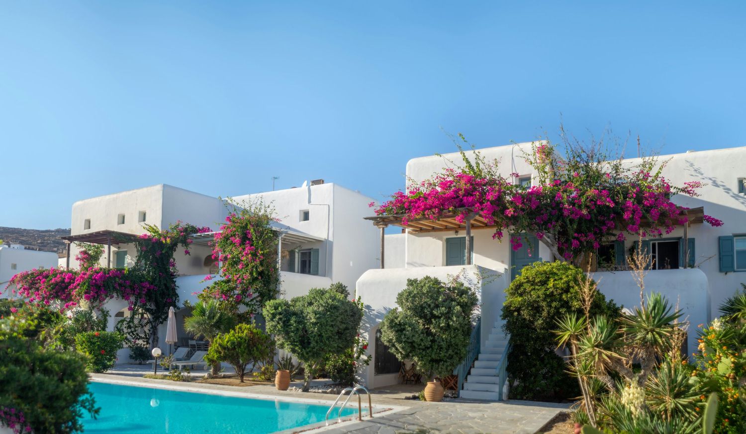 Scenic Mediterranean villa complex with white buildings, swimming pool, and vibrant bougainvillea flowers against blue sky.