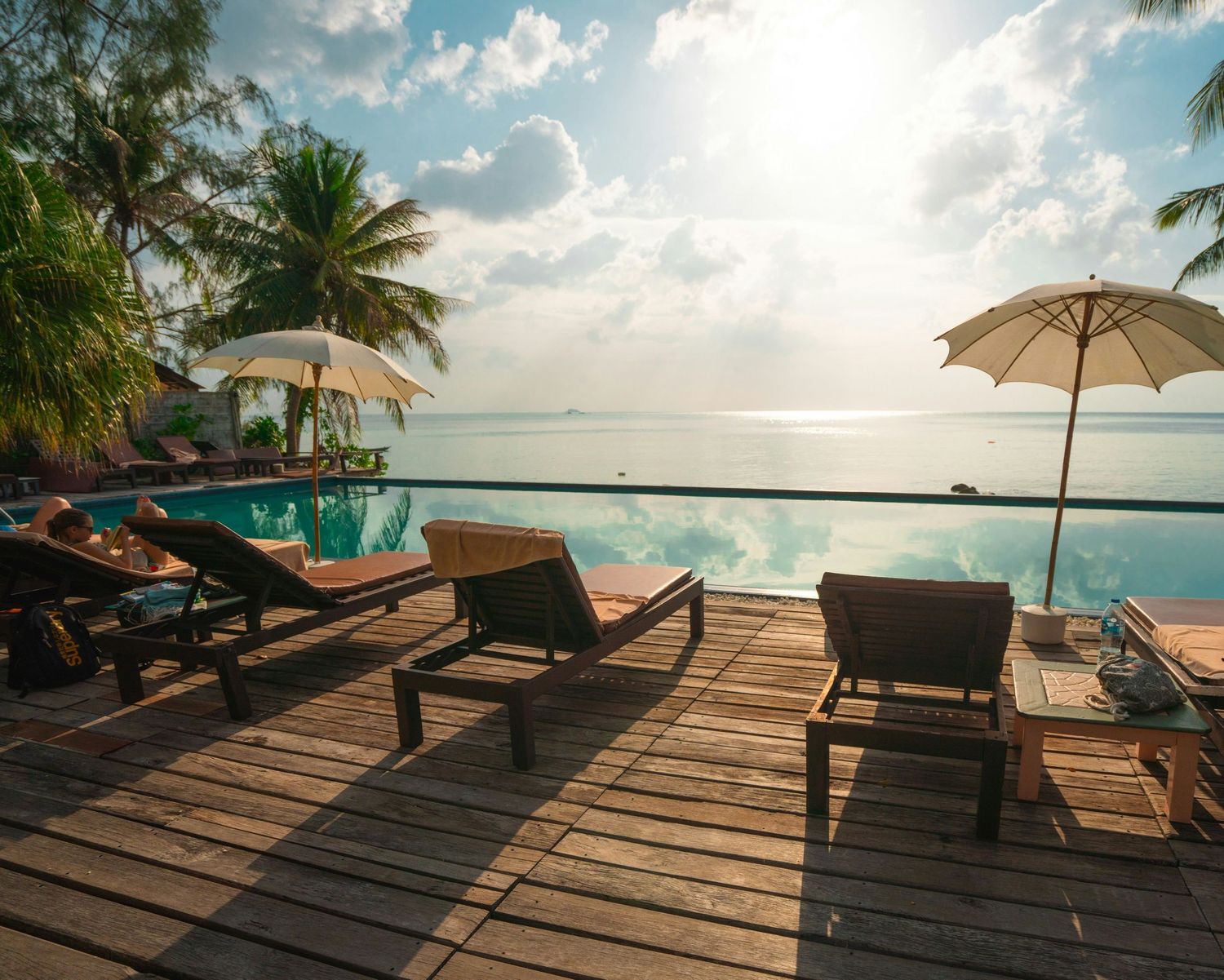 Lounge chairs with umbrellas on a wooden deck overlooking a tropical beach at sunset create a peaceful paradise scene.