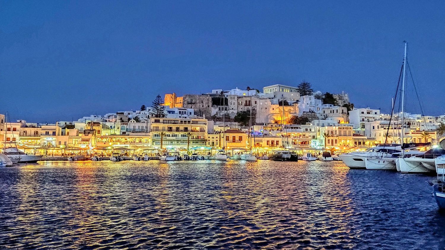 Panoramic view of illuminated coastal Mediterranean town with boats in harbor at dusk showing colorful waterfront buildings.