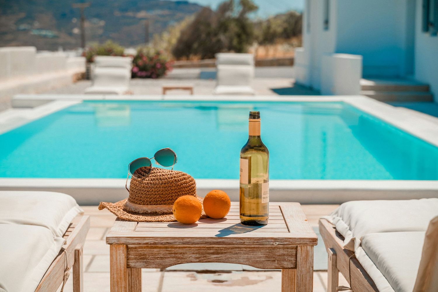 Straw hat, sunglasses, oranges and wine bottle on wooden table beside turquoise swimming pool at luxury resort.