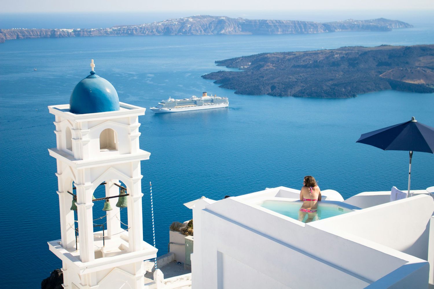 White church bell tower with blue dome overlooking Santorini caldera with cruise ship and Mediterranean Sea views.