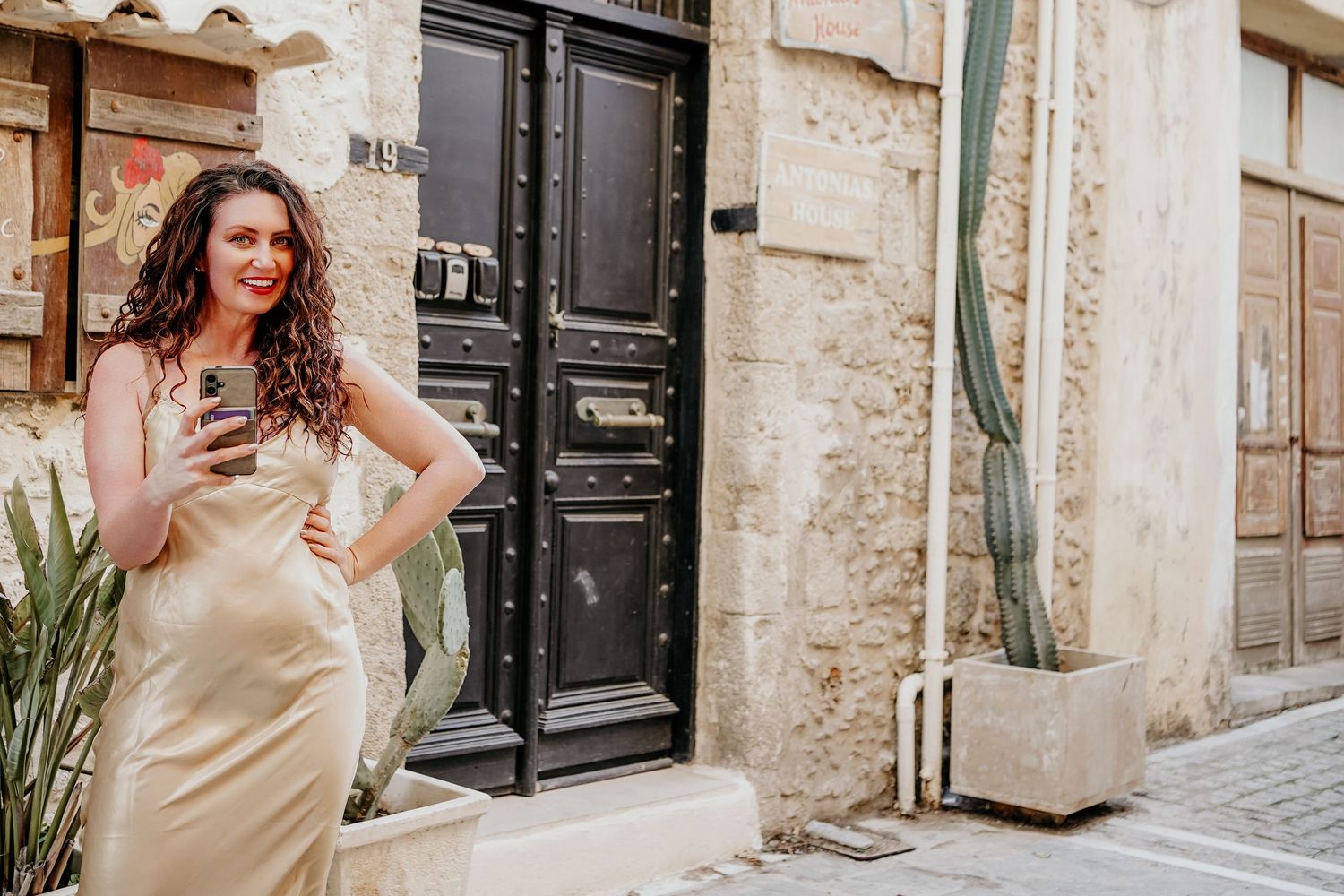 Cristina Nicole Chief Marketing Officer in a beige silk dress poses against a rustic stone wall with black doors in the background.