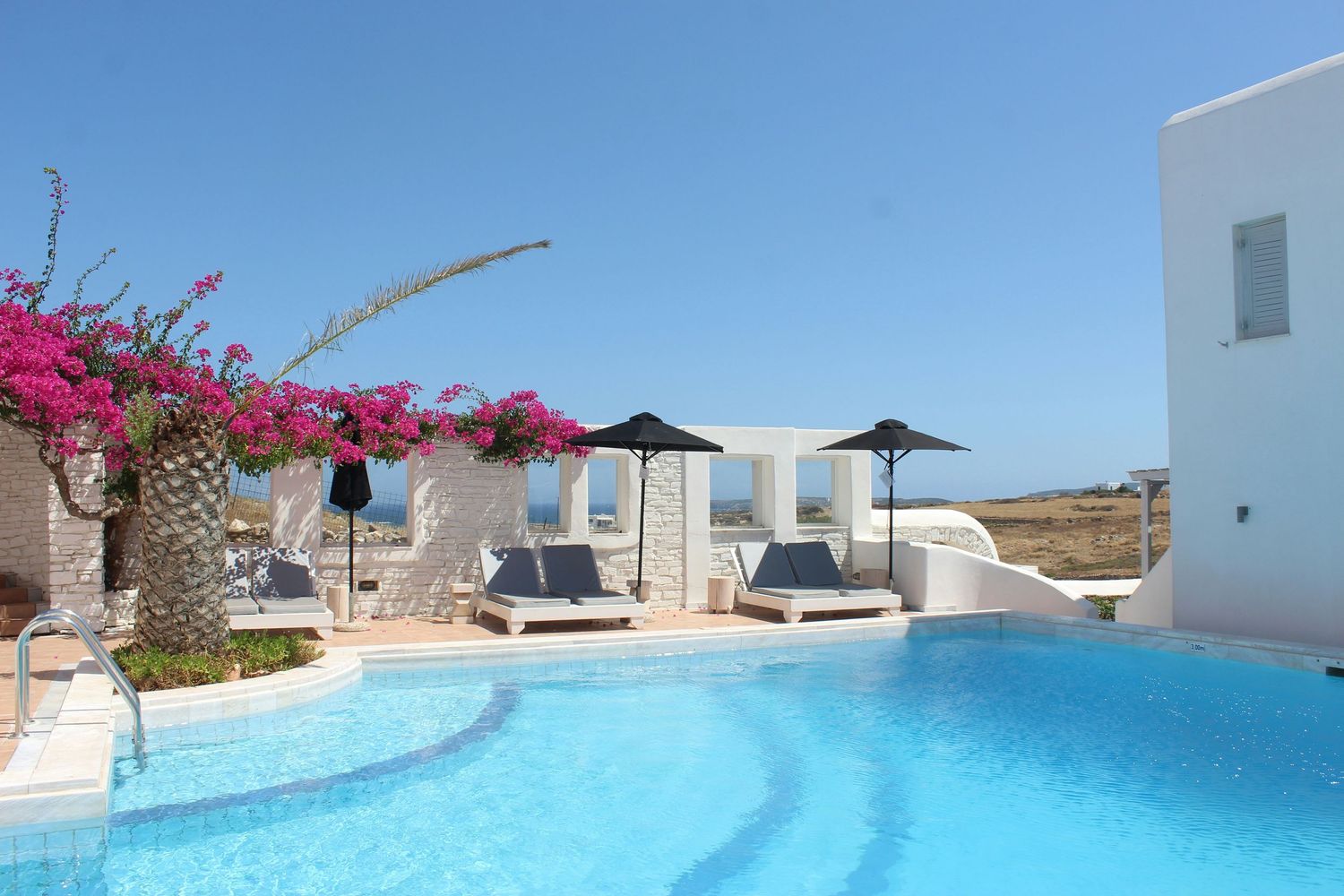 Stunning infinity pool overlooking Mediterranean waters with white architecture and pink bougainvillea flowers in Greece.