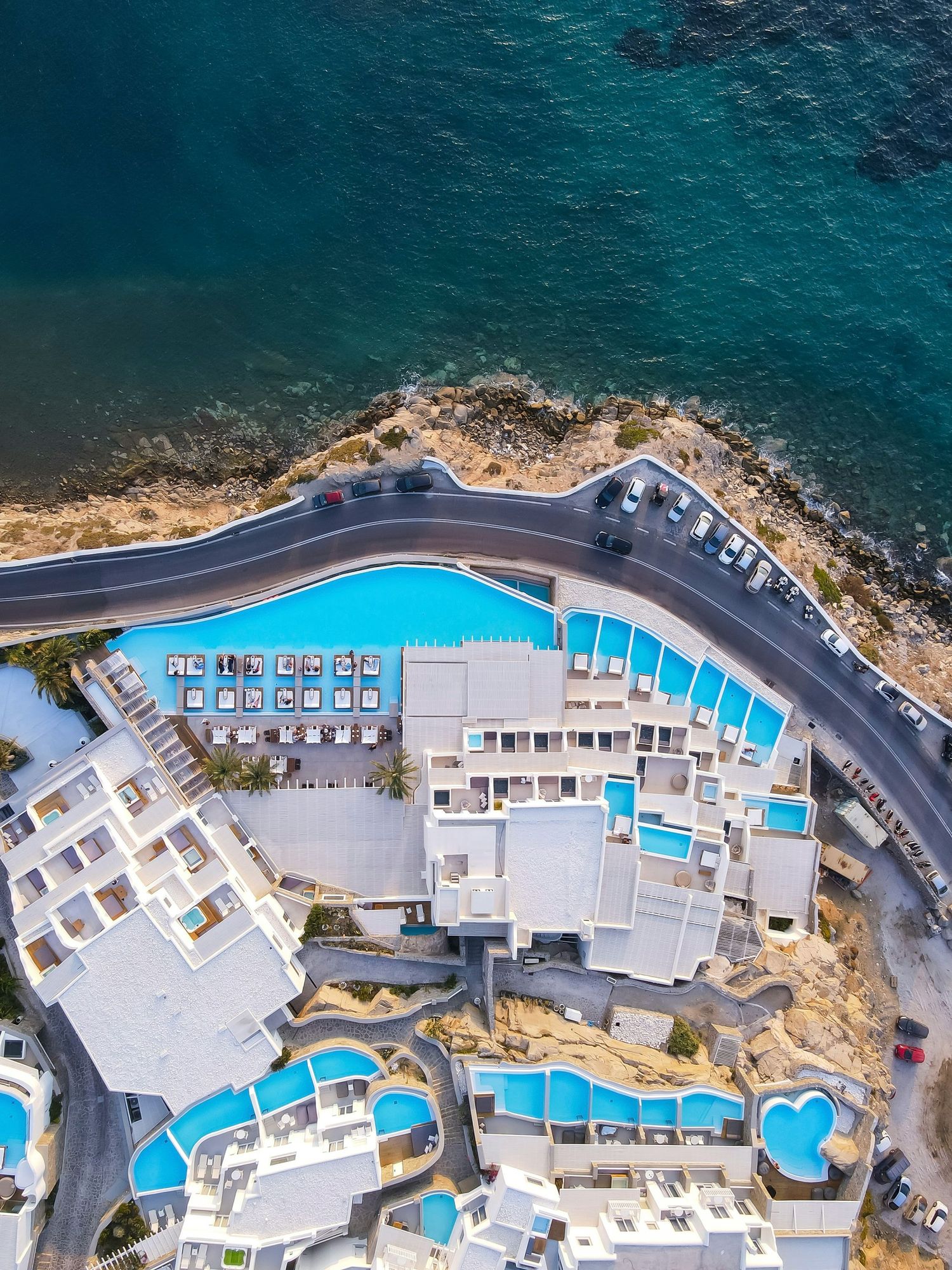 Aerial view of luxury coastal resort with multiple swimming pools and curved road along Mediterranean shoreline.