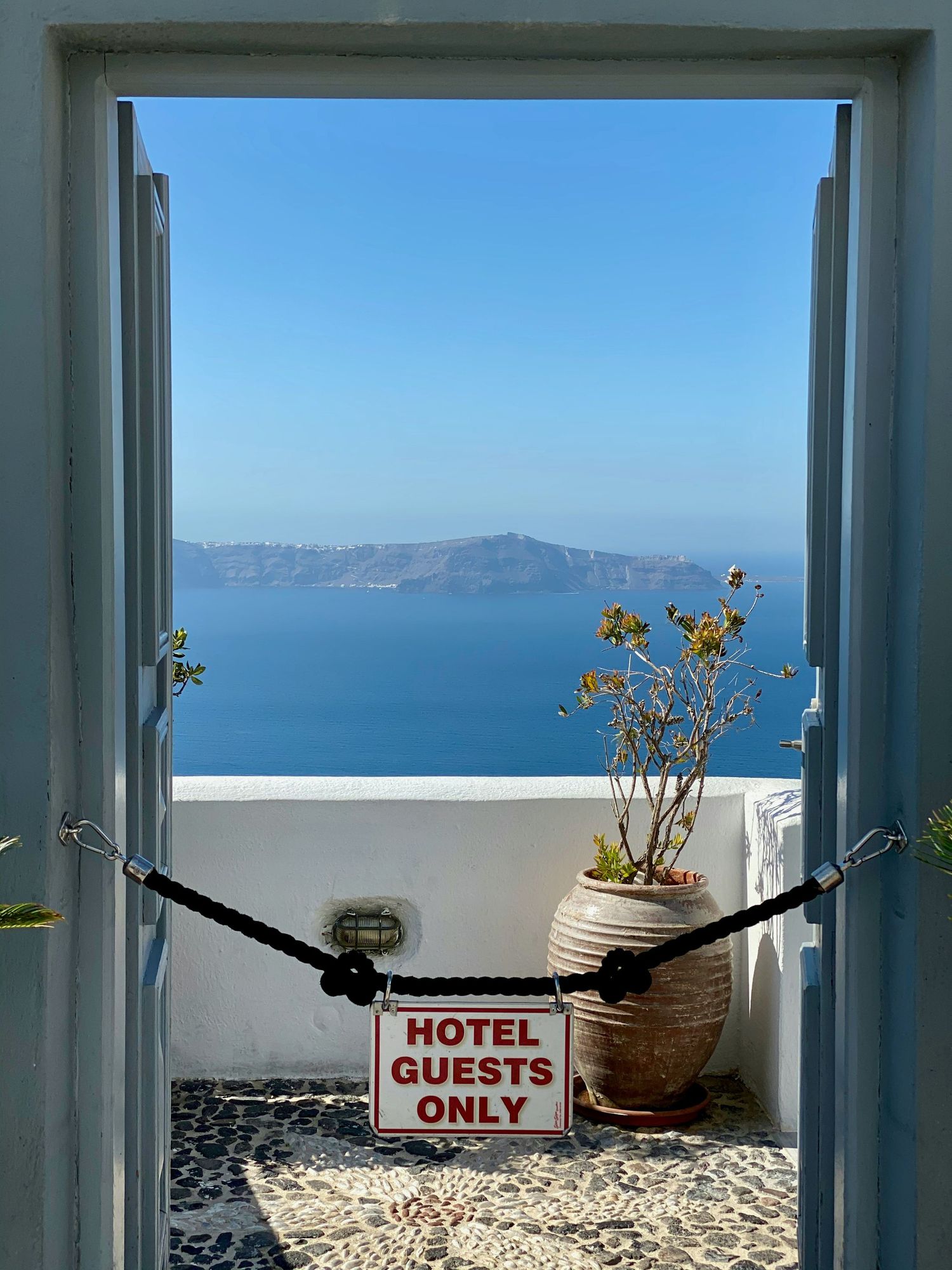 A hammock with ocean view through white-framed windows in Santorini, with 'Hotel Guests Only' sign and potted plants.