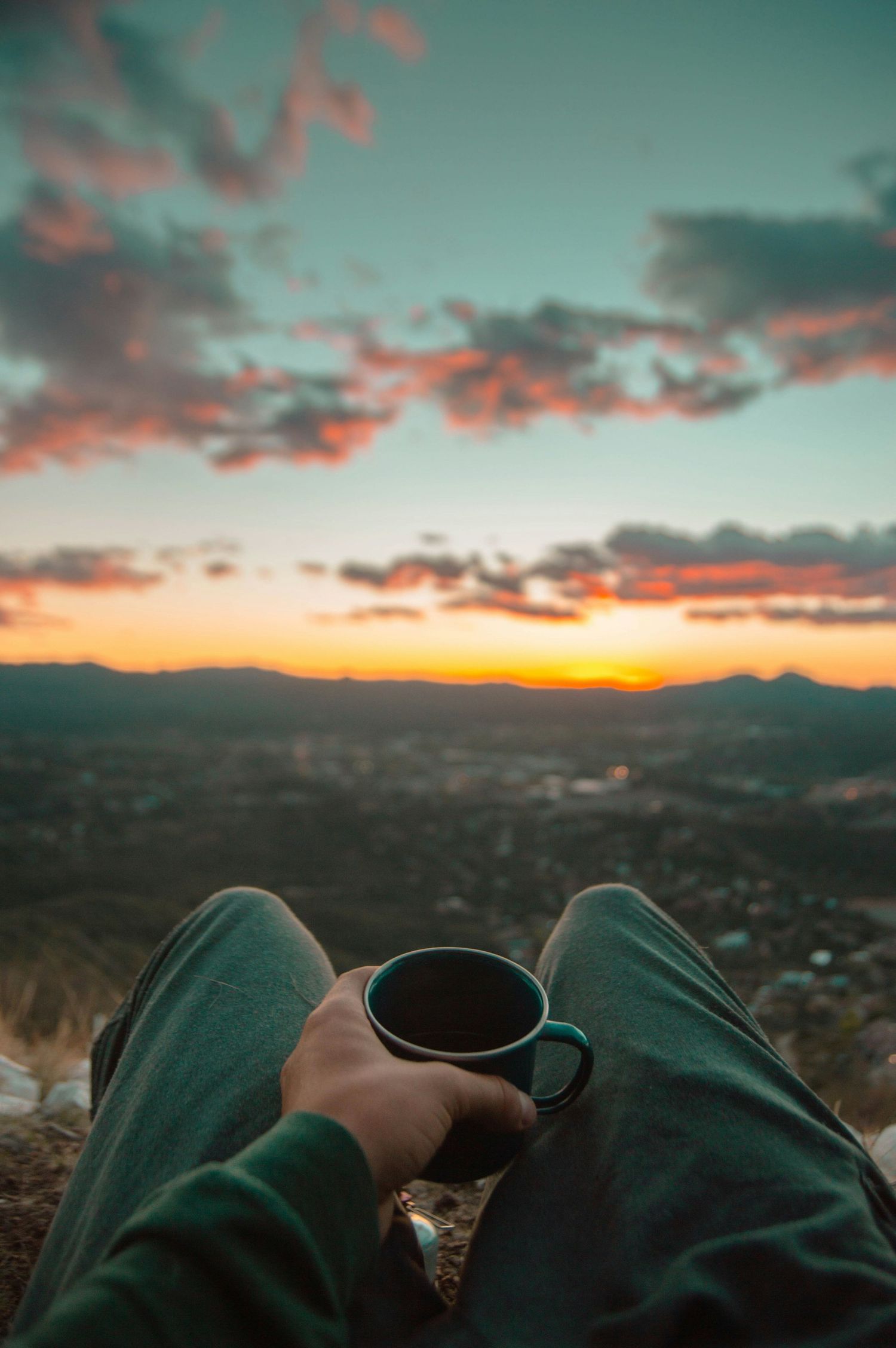 Person holding coffee mug while enjoying scenic mountain sunset view in green pants with clouds in orange sky.