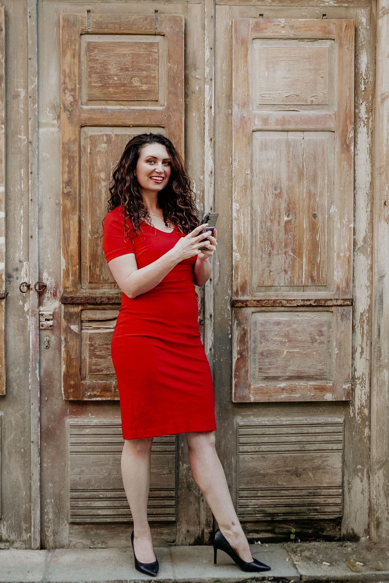 Cristina Nicole, Chief Marketing Officer in a fitted red dress and black heels leaning against weathered wooden doors while holding a phone.