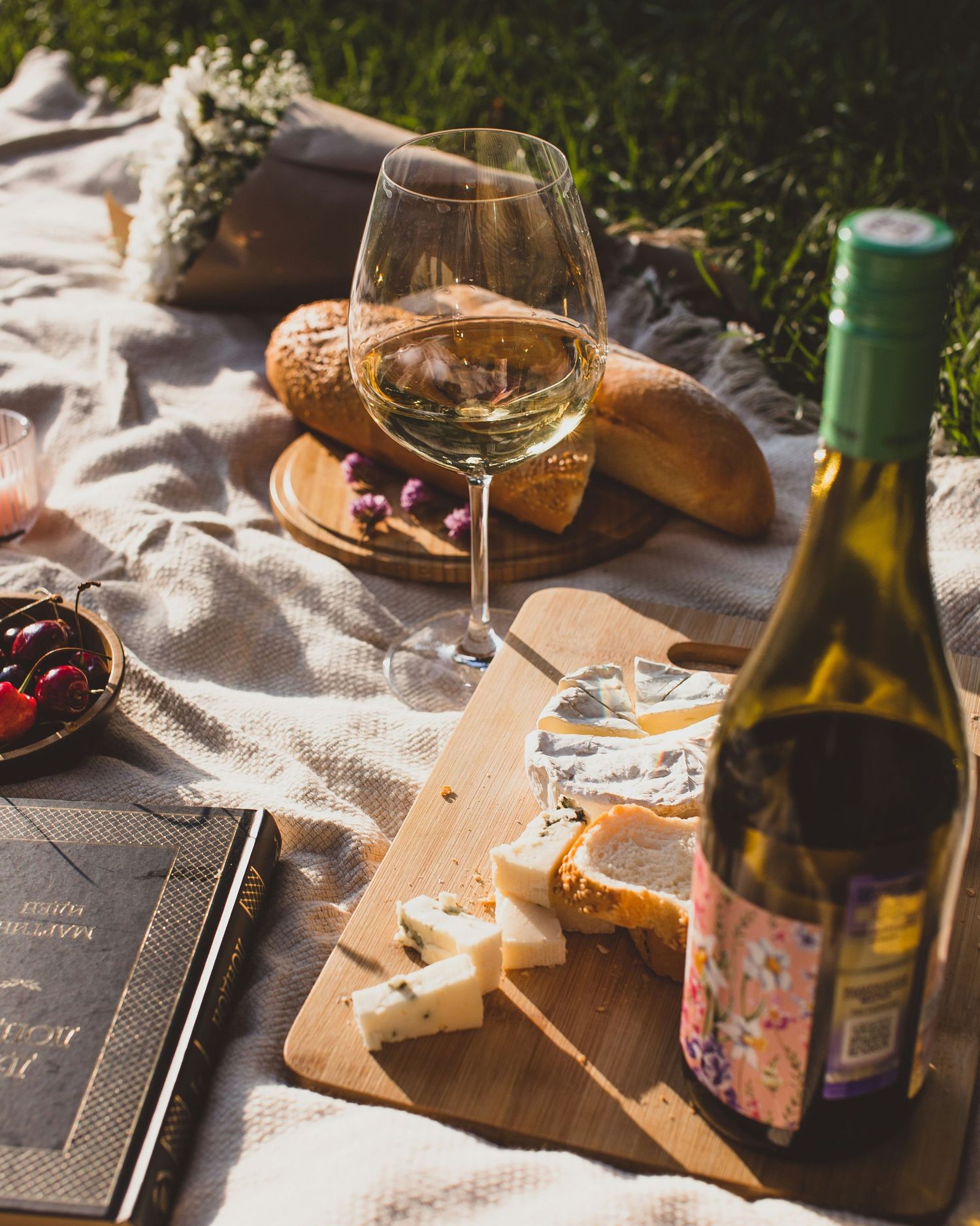 A rustic picnic scene with wine glasses, cheese boards, and bottles arranged on a textured tablecloth in natural sunlight.