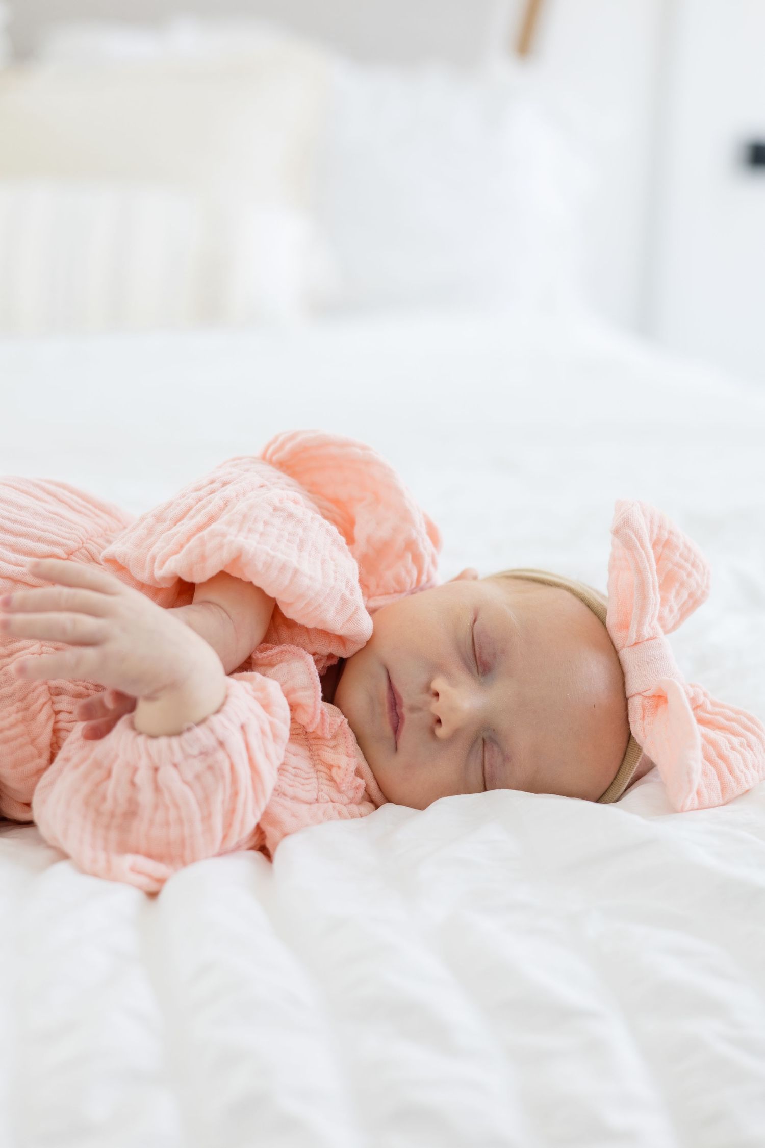 Newborn baby girl in pink outfit and bow on white bed during lifestyle newborn session in the summer with McKinney newborn photographer