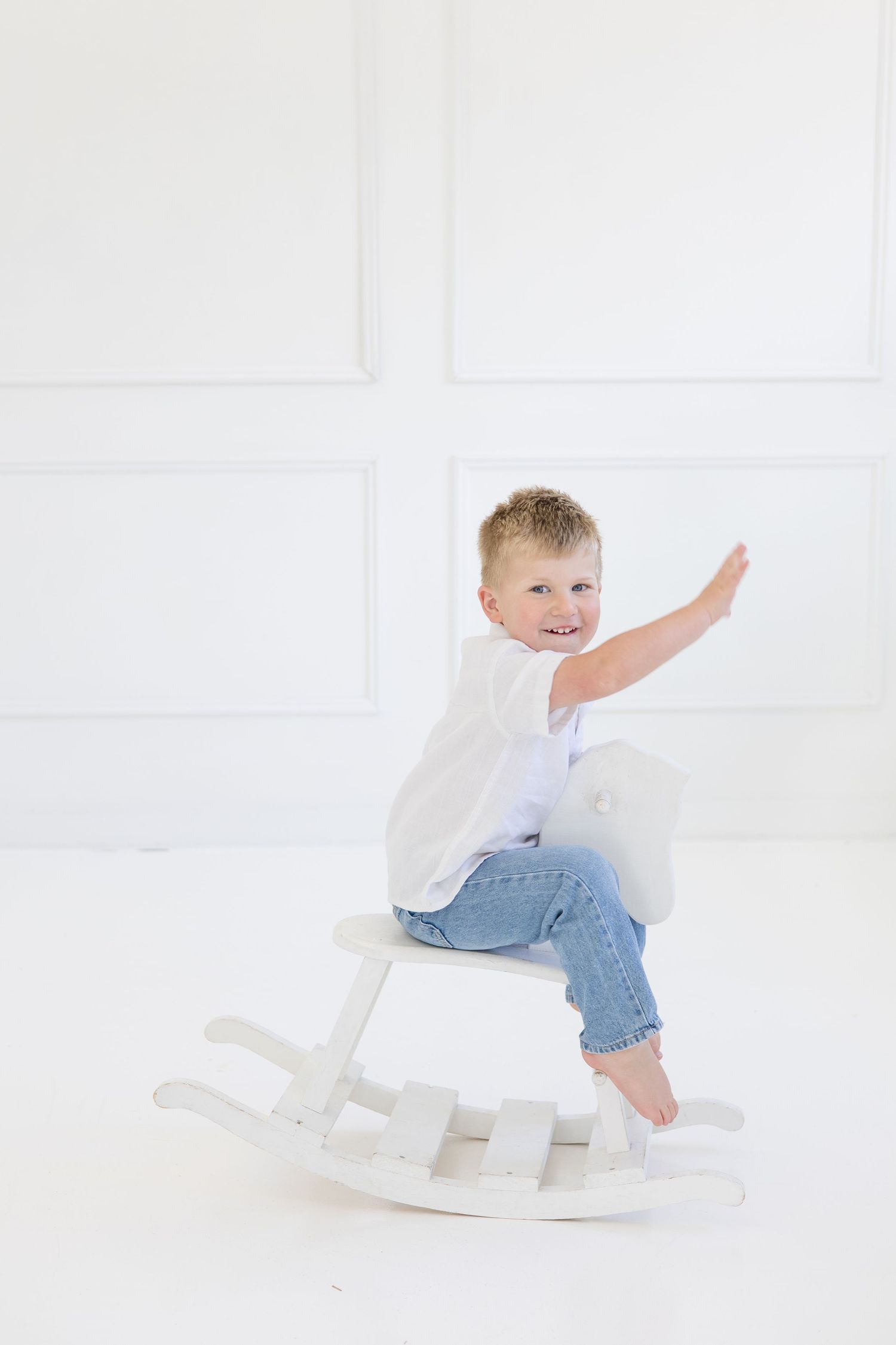 Big brother plays rodeo on rocking horse in all-white studio during family lifestyle newborn session in the summer McKinney photographer