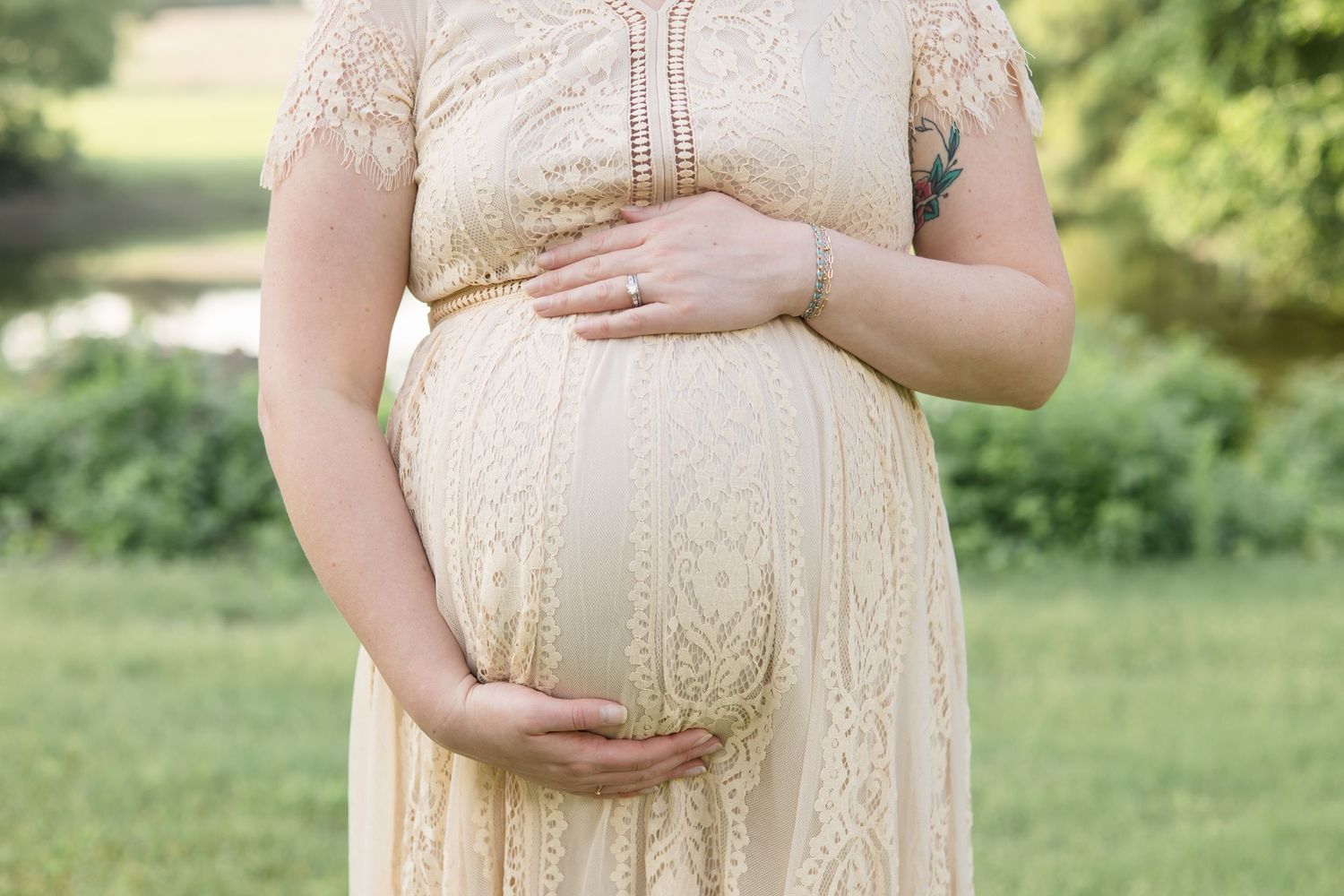 expecting mom in tan cream dress holds belly in front of pond at erwin park during maternity session with mckinney maternity photographer