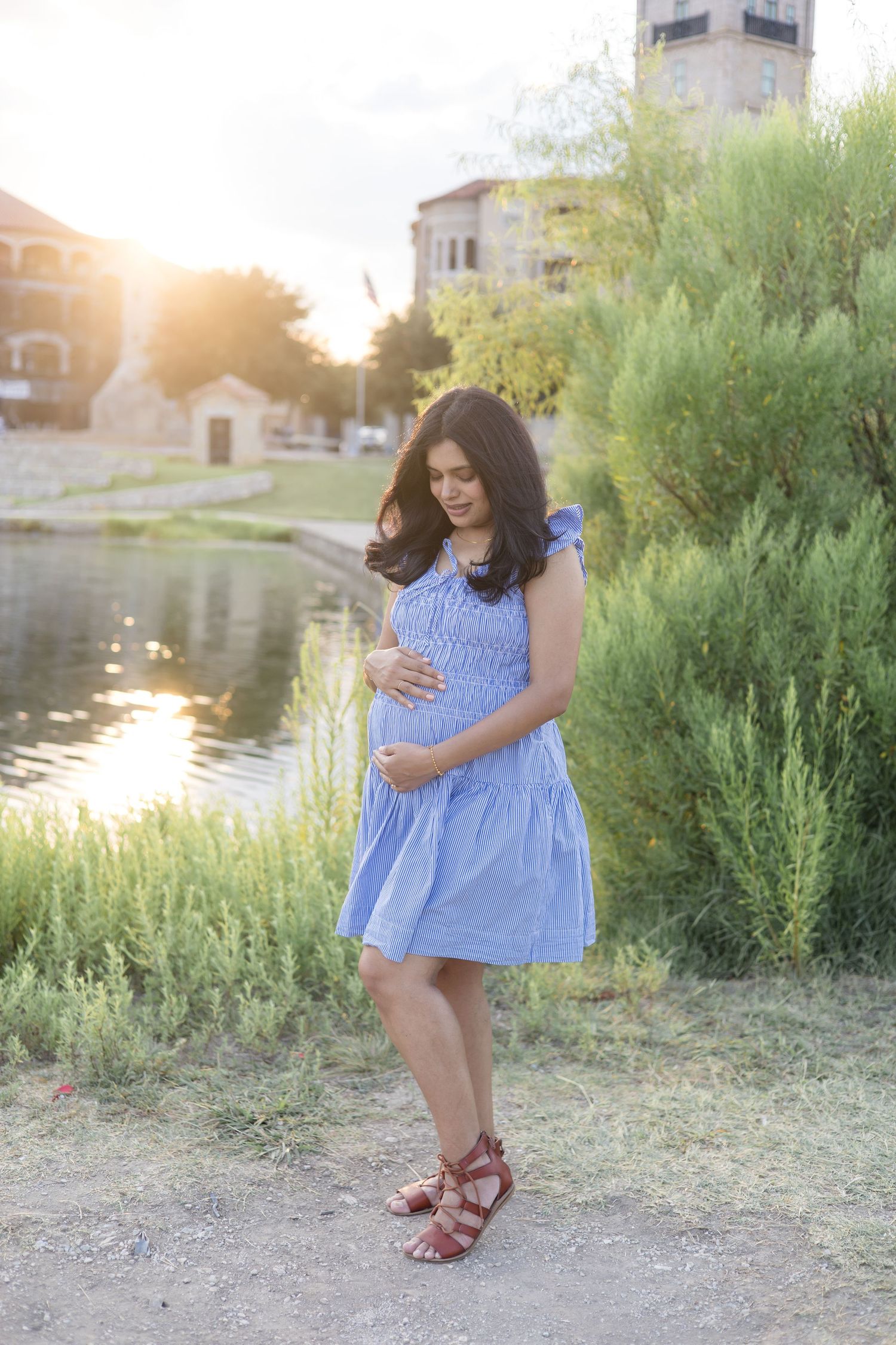 mom holds pregnant belly in blue dress during maternity session with McKinney Family Photographer in the fall at Adriatica Village