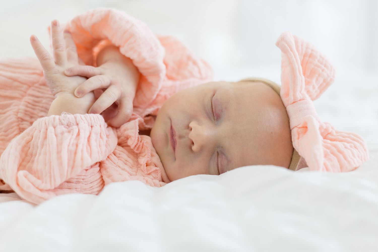 Newborn baby girl in pink outfit and bow on white bed during lifestyle newborn session in the summer with McKinney newborn photographer
