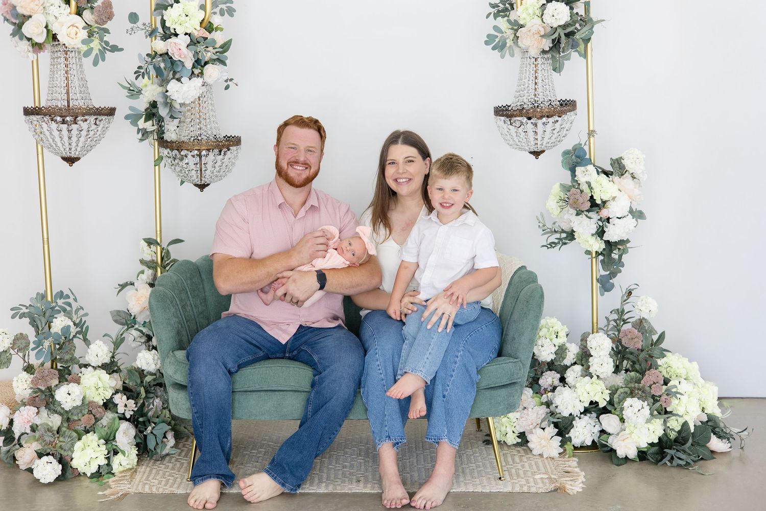 Family in jeans, white and pink pose together on green couch in front of floral chandelier set at Lemon Drop Studio for lifestyle newborn