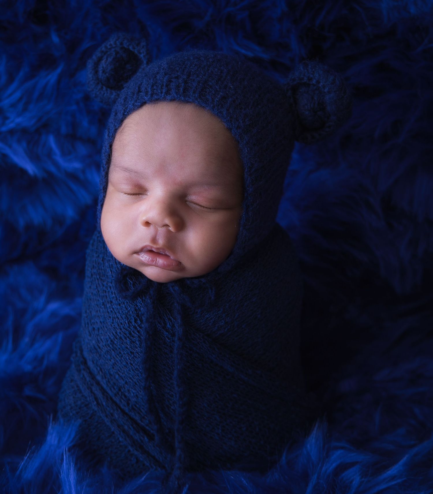 Sleeping newborn wrapped in dark blue blanket against deep blue textured background.