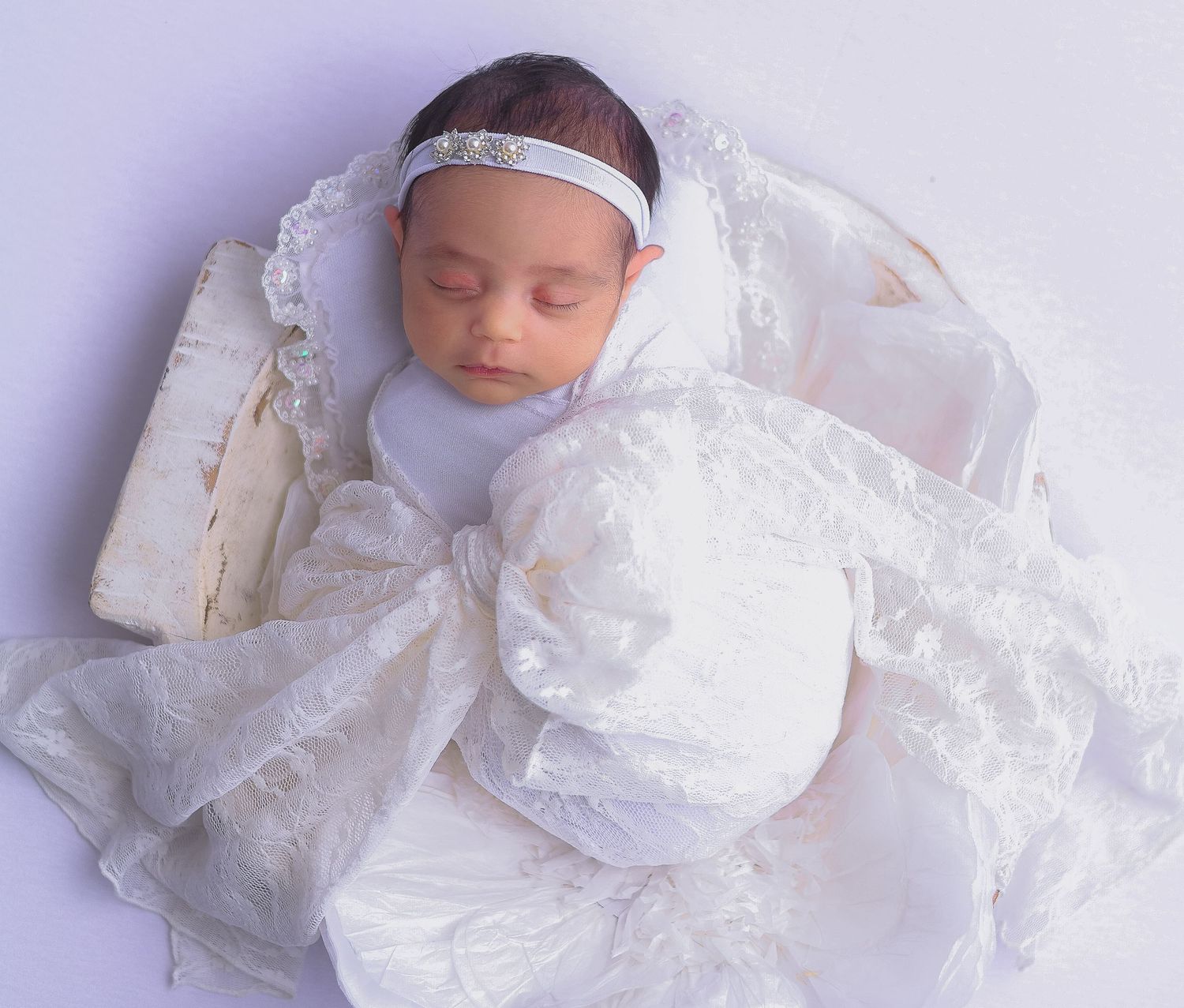A sleeping newborn baby wearing an elegant white christening gown and headband rests peacefully on white bedding.