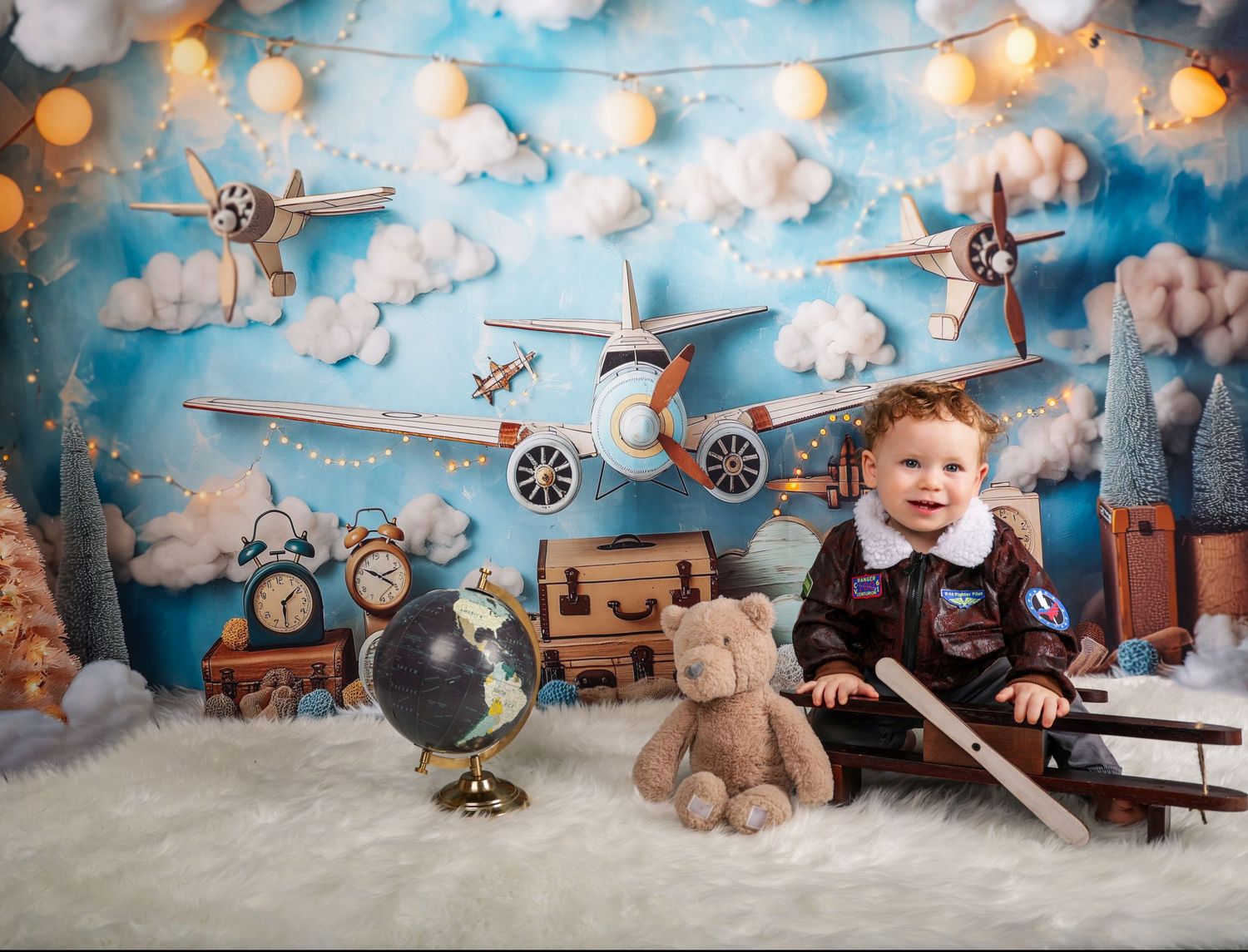 Vintage aviation themed setup with toy airplanes, teddy bear, and string lights against cloudy blue backdrop.