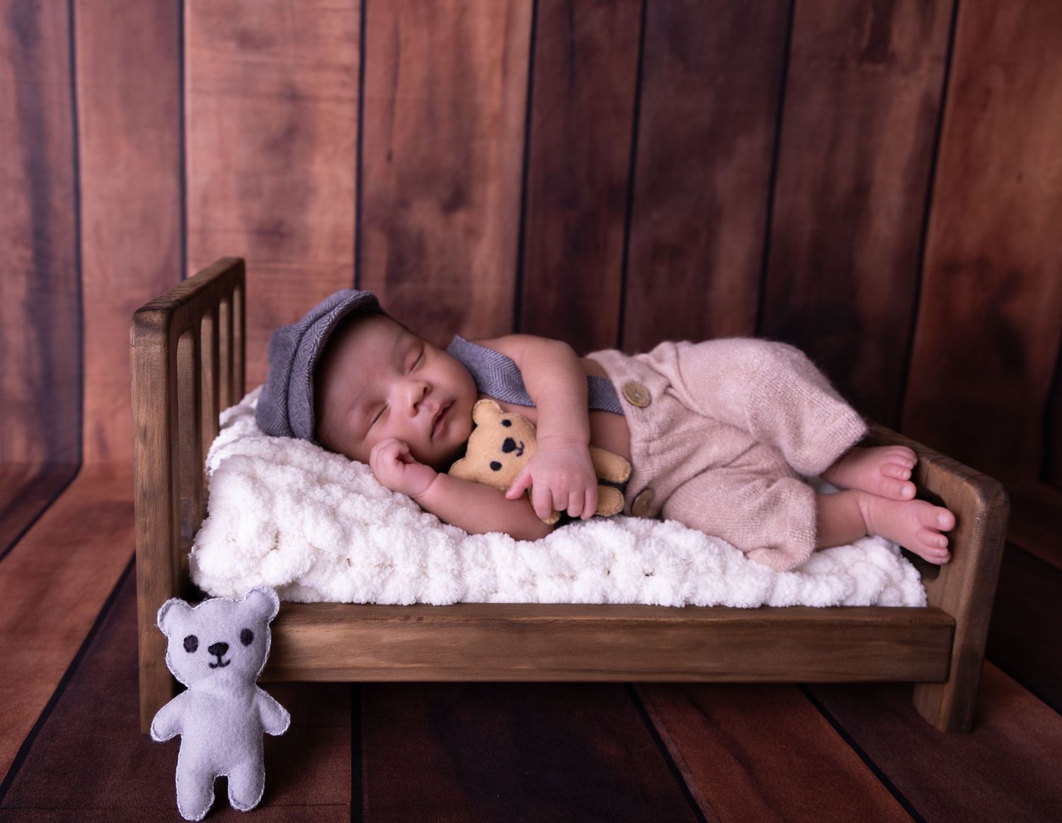 Baby in gray cap sleeping peacefully on white blanket with plush teddy bears on wooden bed.