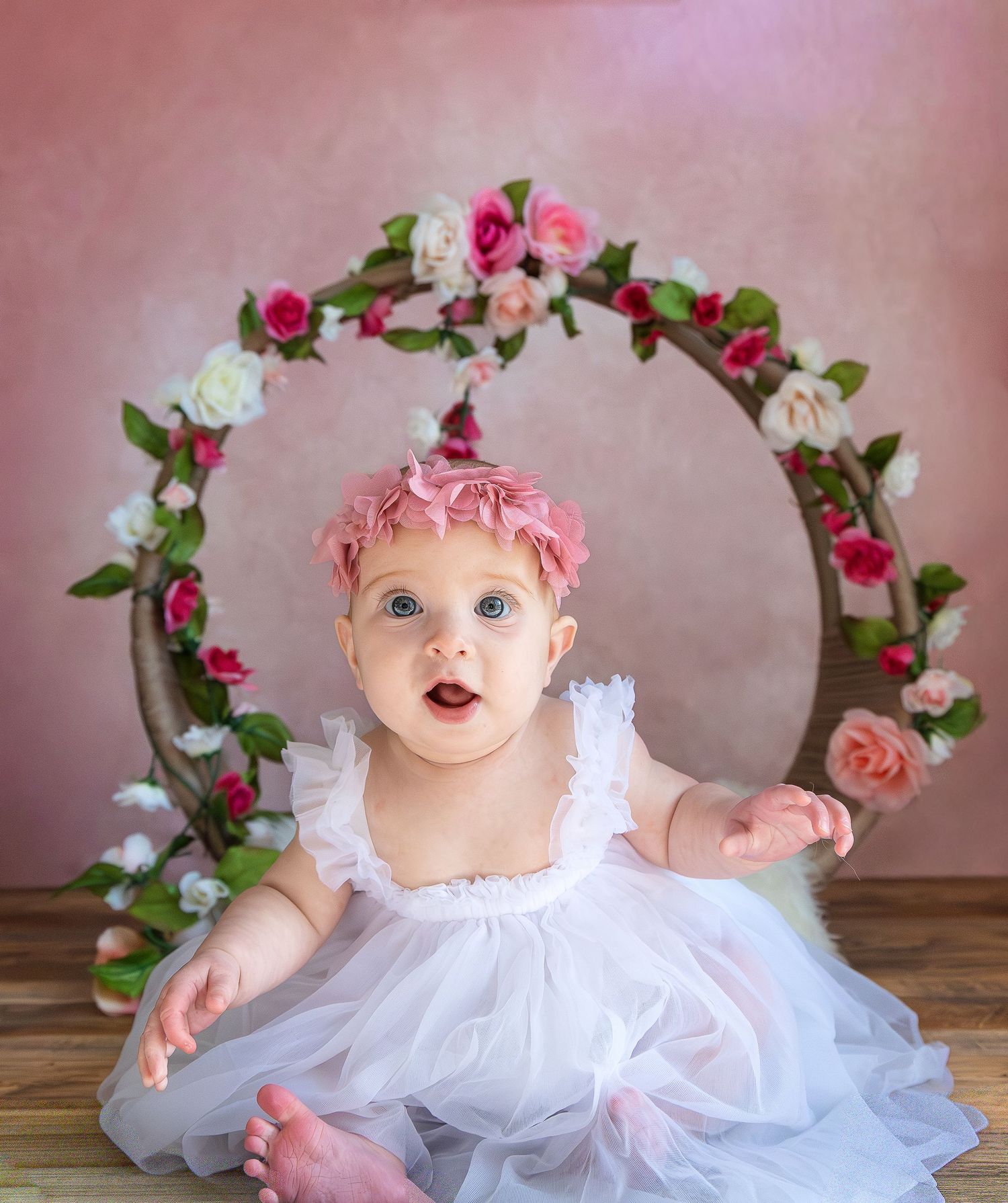 A baby in a white tulle dress and pink floral crown poses for a series of professional photographs on a wooden floor.