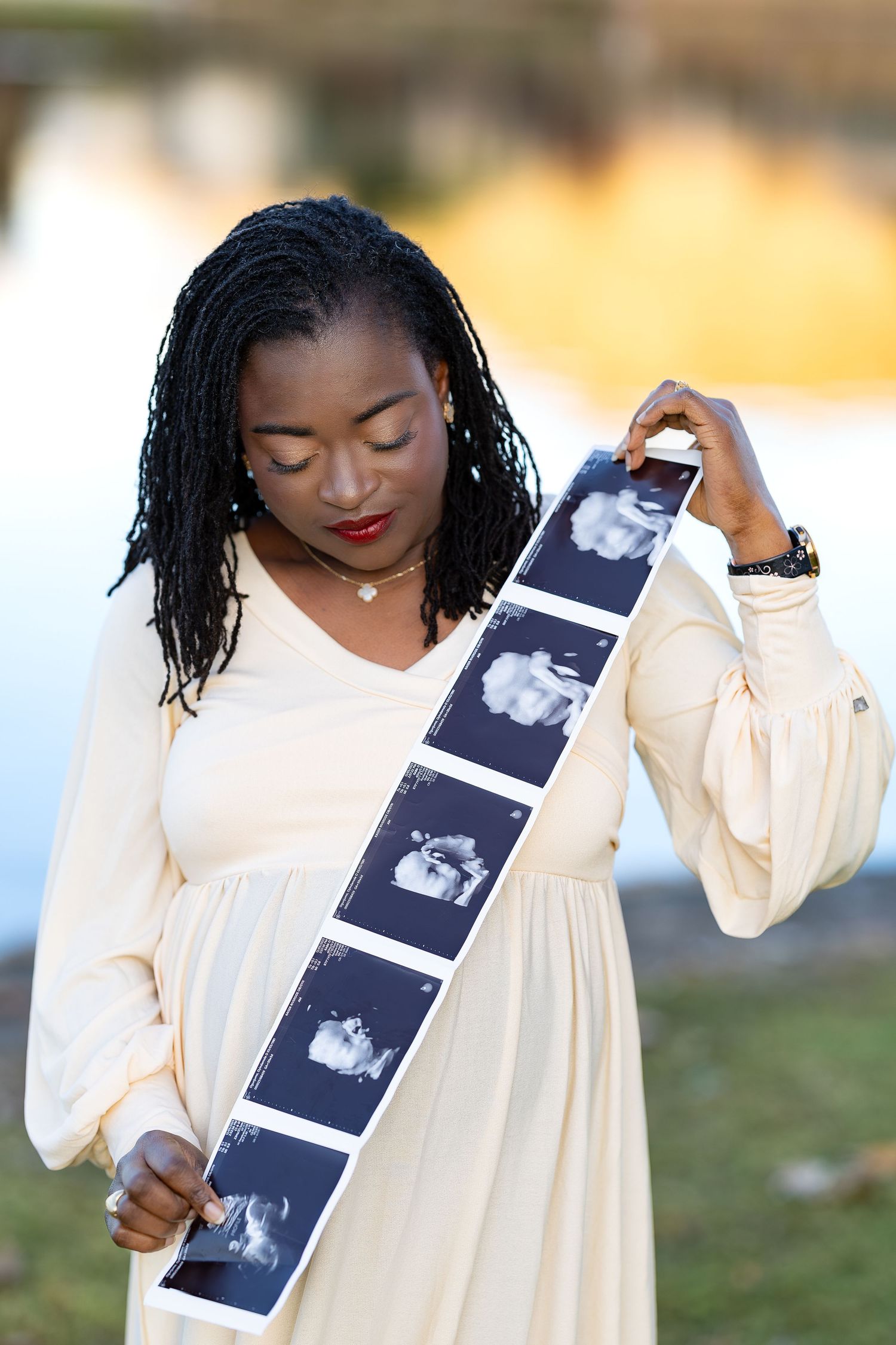 A person in a flowing white dress holds up a strip of ultrasound photos while posing outdoors.